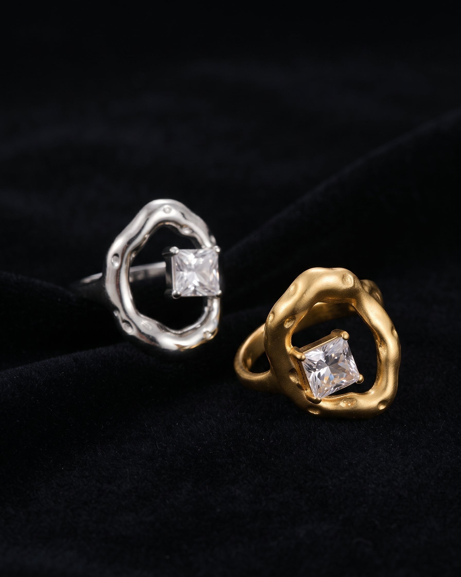 Two abstract ring earrings displayed on a black velvet background, one in silver and one in gold, each featuring a princess-cut clear gemstone.