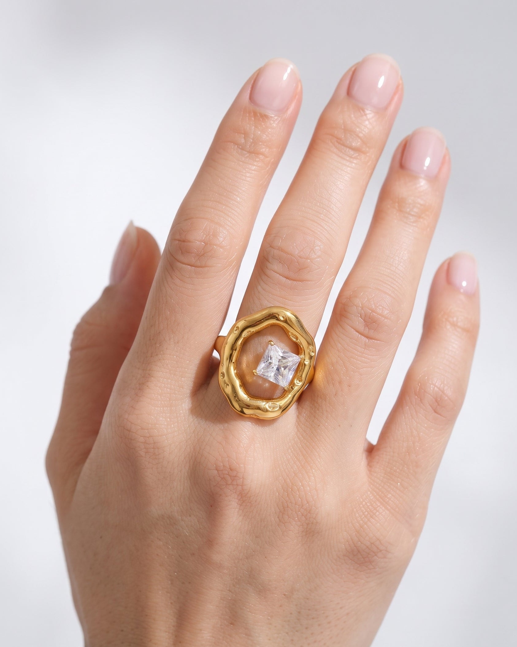 A close-up of a hand wearing a textured gold ring with a clear square-cut gemstone, set against a soft light gray background.