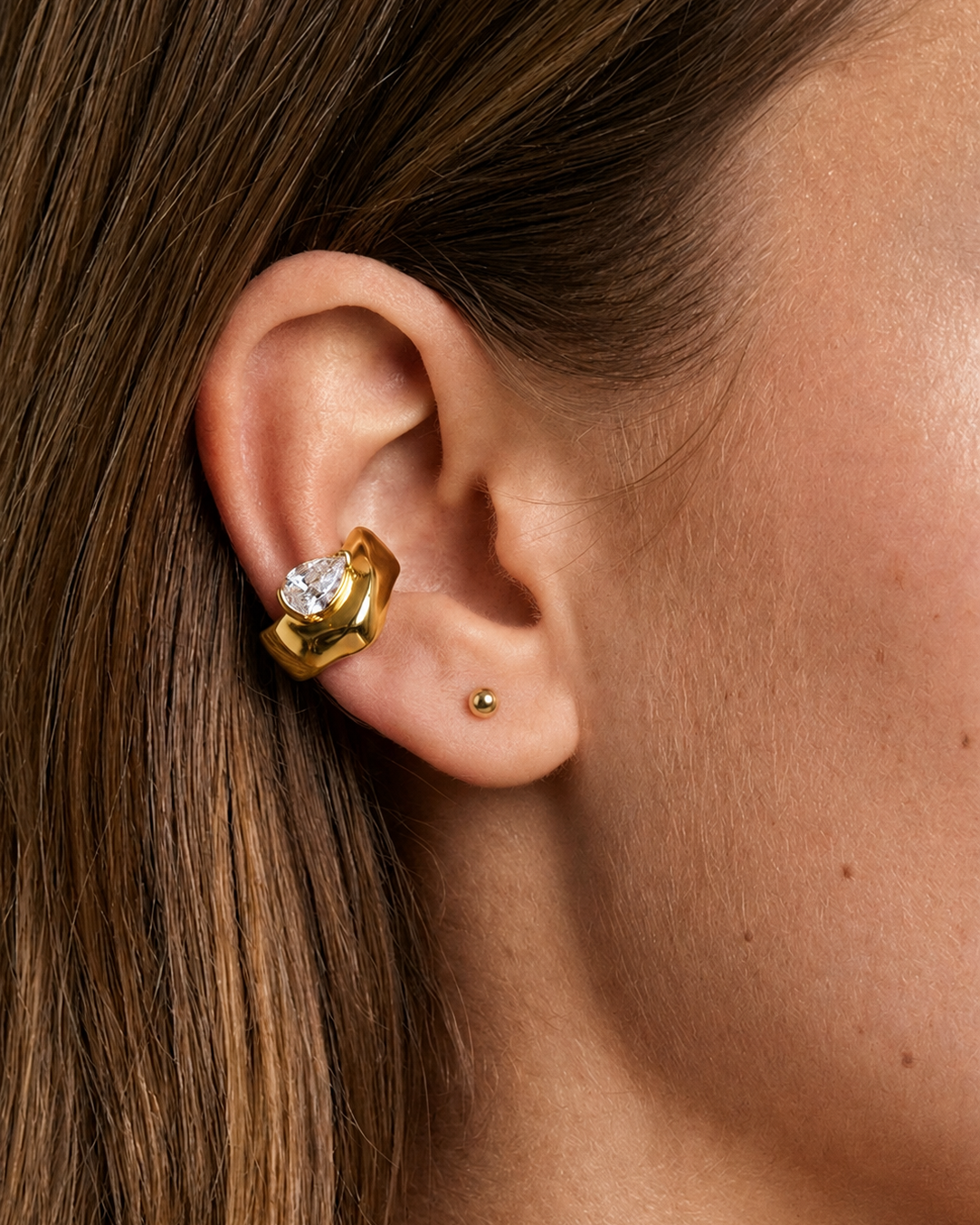 A close-up of a person's ear wearing a gold huggie hoop earring with a clear heart-shaped gemstone, alongside a small gold stud earring.