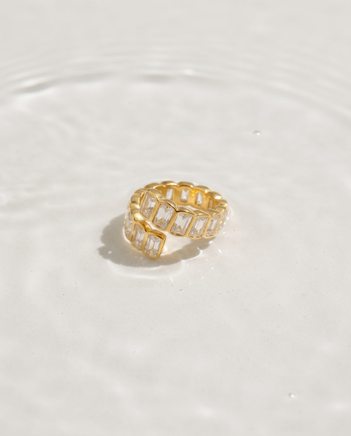 A gold band ring featuring a series of clear baguette-cut stones, photographed on a light off-white surface with soft water ripples.