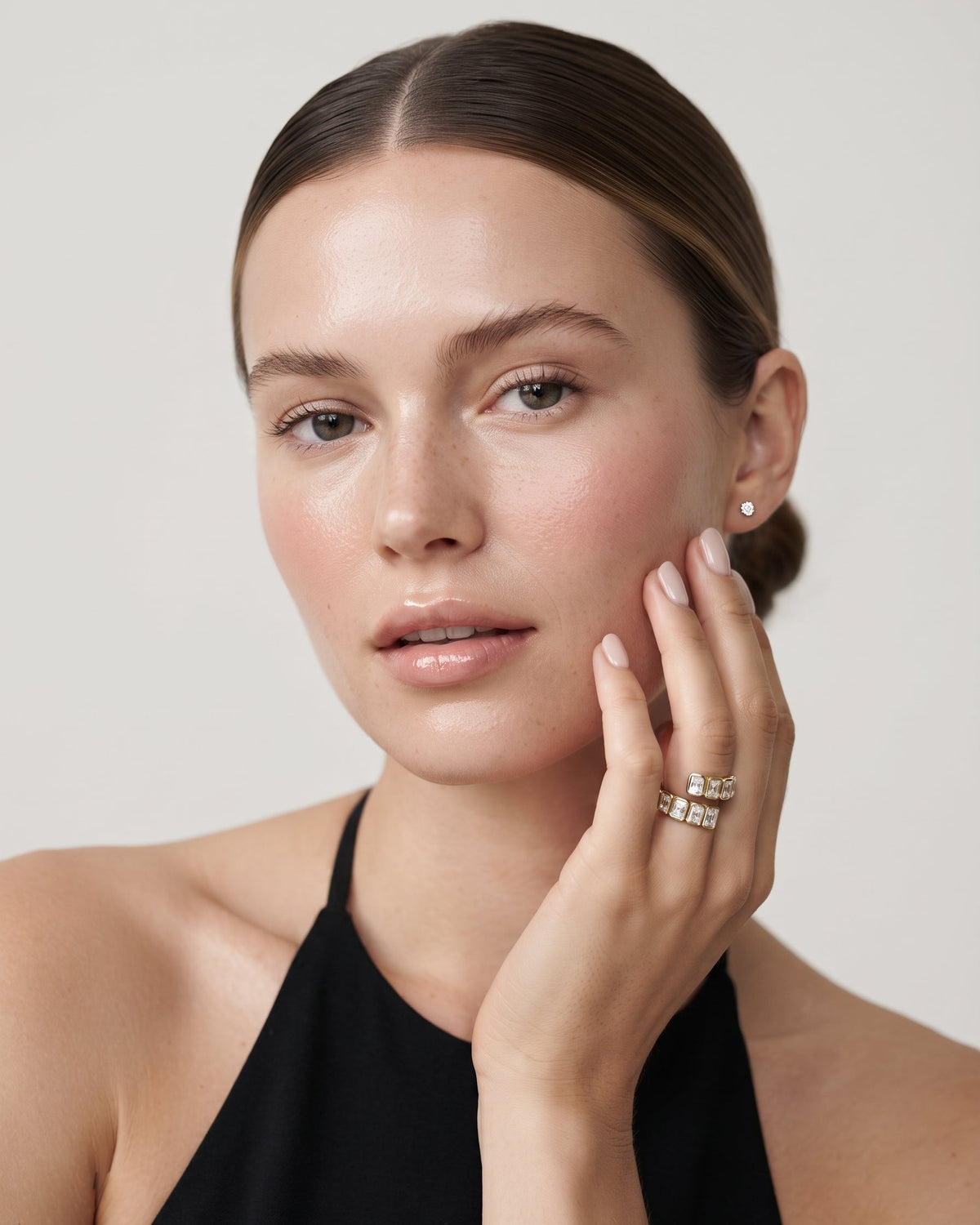 A close-up portrait of a woman with her hair pulled back, wearing a black halter top and delicate silver rings on her fingers, set against a solid light gray background.