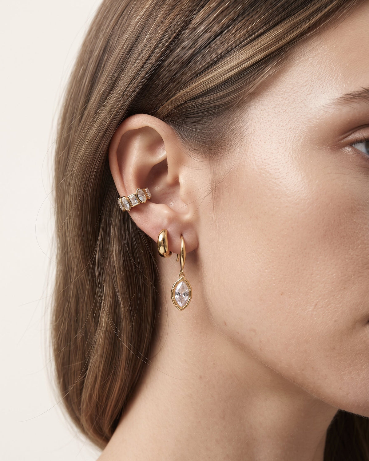 Close-up of a model's ear showcasing a stack of gold ear cuff and drop earrings with clear marquise-cut gemstones against a light beige background.