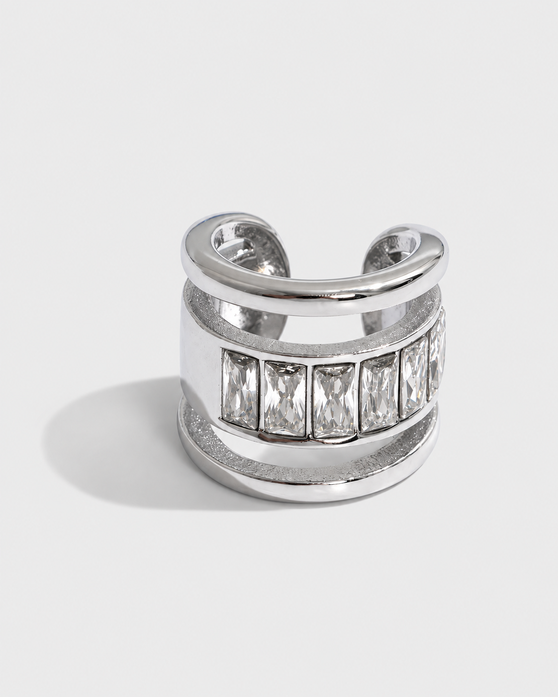 A wide silver-toned cuff ring featuring a row of clear rectangular baguette gemstones on the front, photographed on a light gray background.