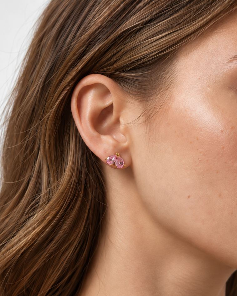 A close-up of a woman's ear wearing a small pink stud earring with multiple clear stones, shown against a light neutral background.