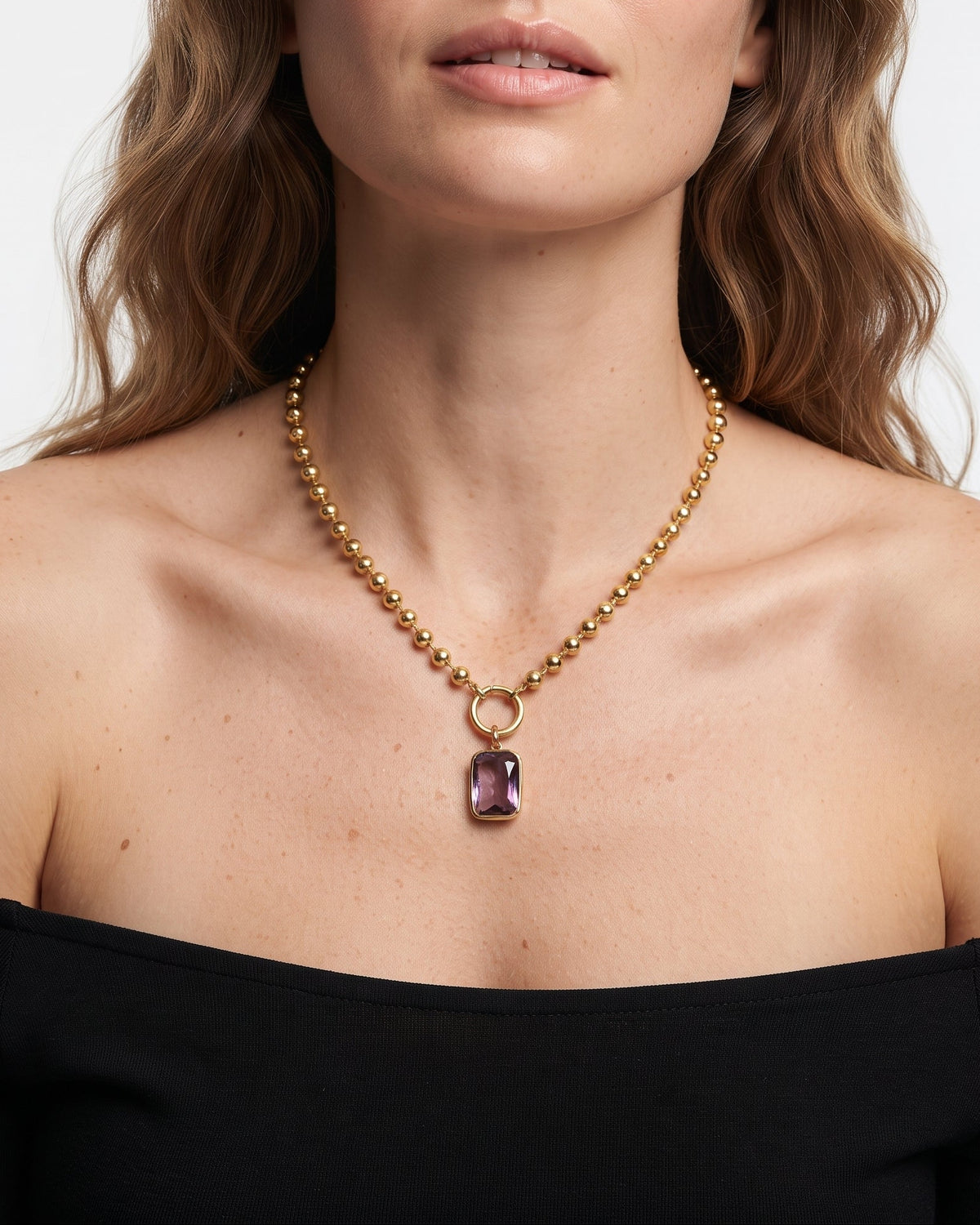 A woman wearing a gold beaded necklace with a rectangular purple gemstone pendant, shown in a close-up portrait against a light gray background.