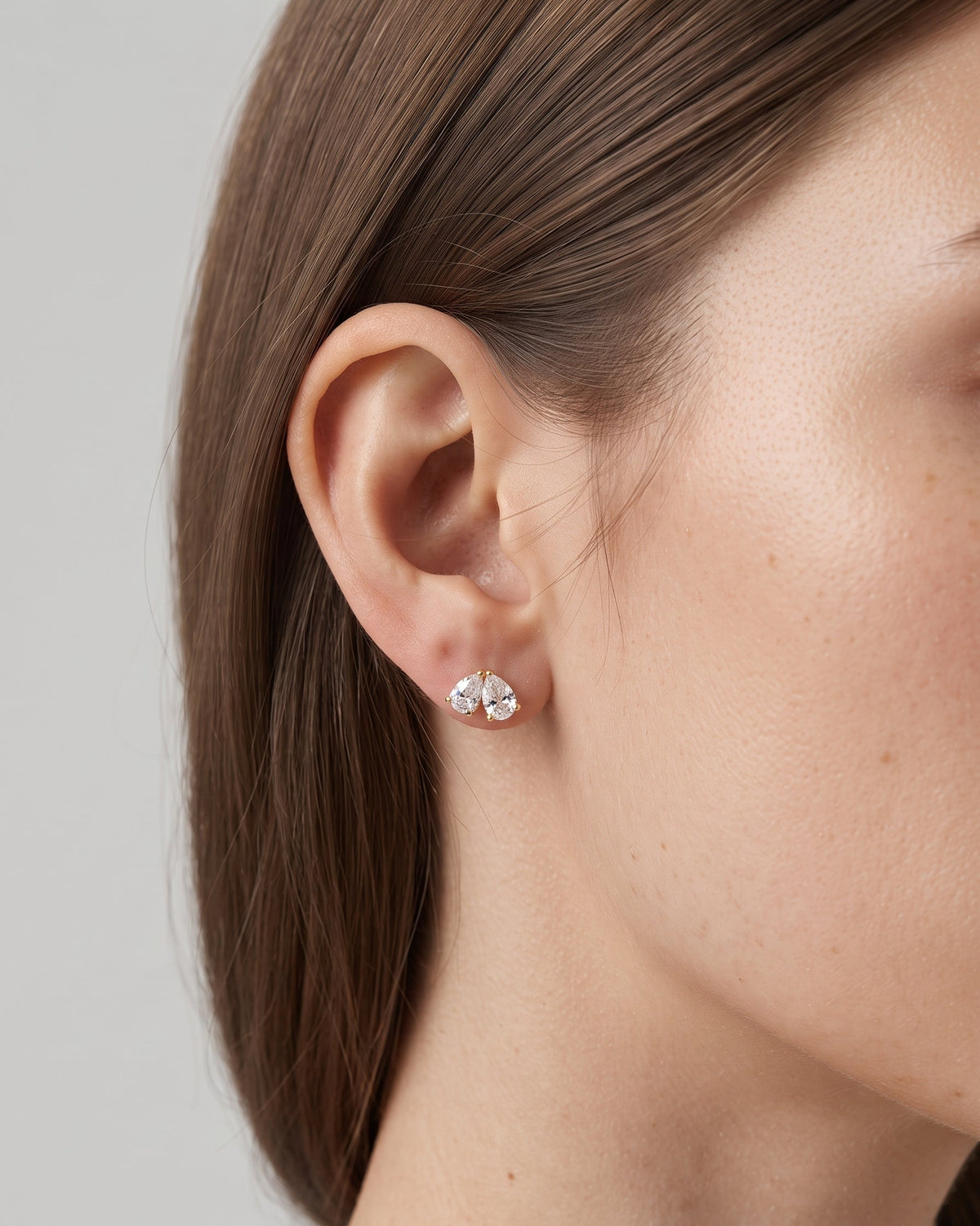 A close-up side profile of a woman's ear wearing a small silver stud earring with two clear heart-shaped gemstones, set against a neutral light gray background.