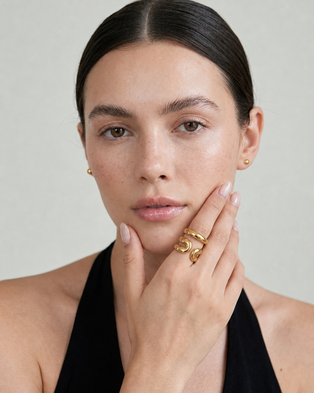 A close-up portrait of a woman with dark hair pulled back, wearing small gold stud earrings and layered gold rings on her fingers, against a light neutral background.