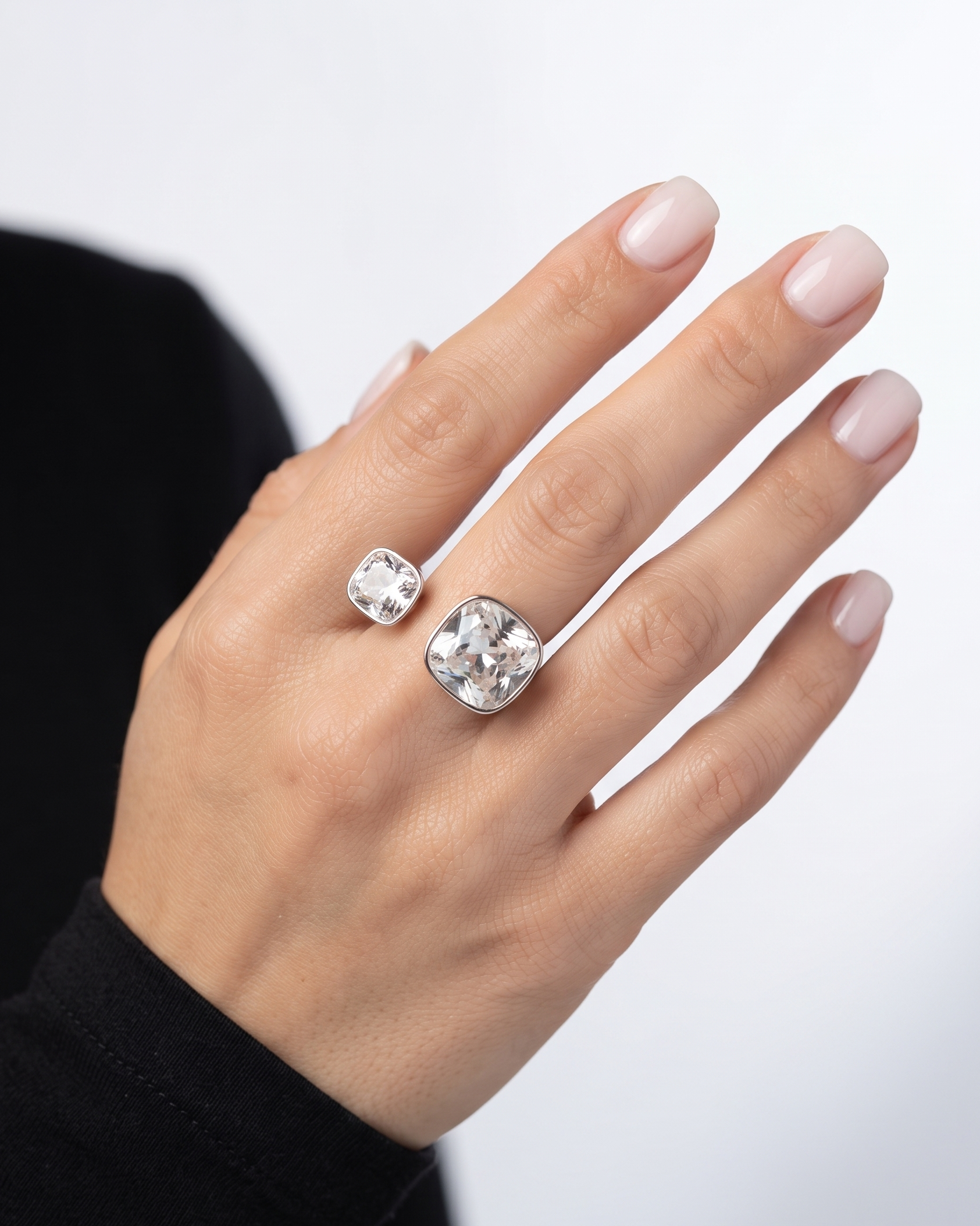 A close-up of a hand wearing two silver-toned rings, including a larger cushion-cut ring and a smaller round ring, against a light gray background.
