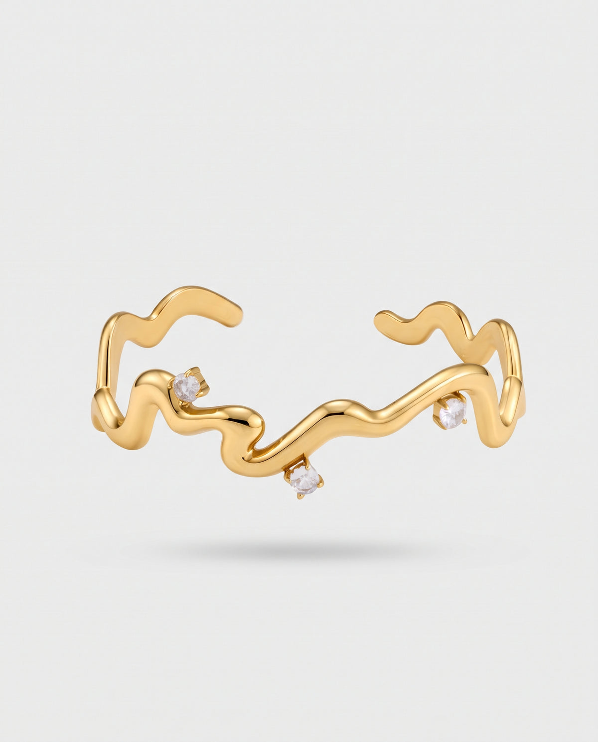 A gold open cuff ring with a wavy, fluid line design and three small clear gemstone accents, shown on a light gray background.