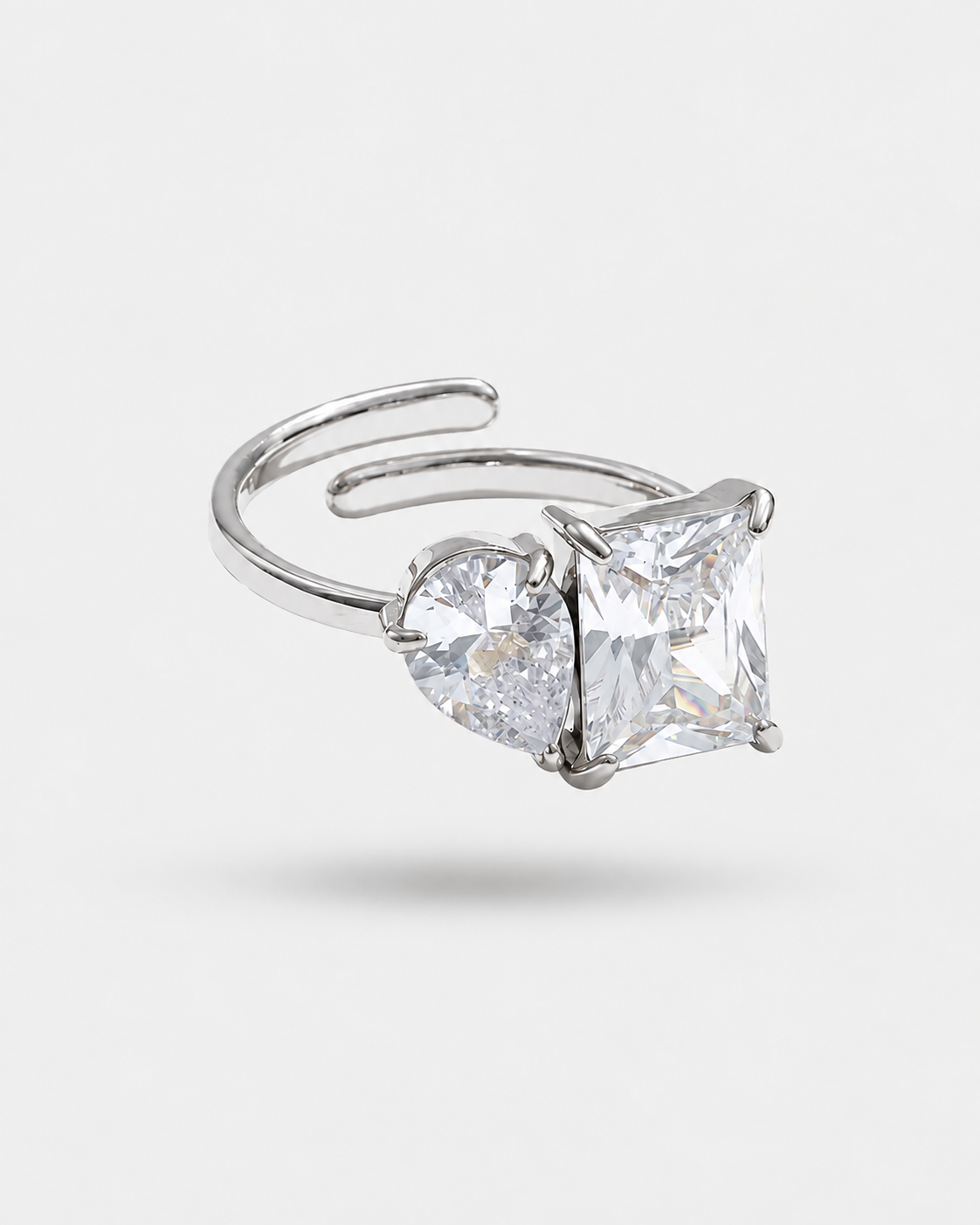 A silver-toned adjustable ring featuring a large princess-cut clear gemstone and a smaller heart-shaped clear gemstone, shown on a light gray background.