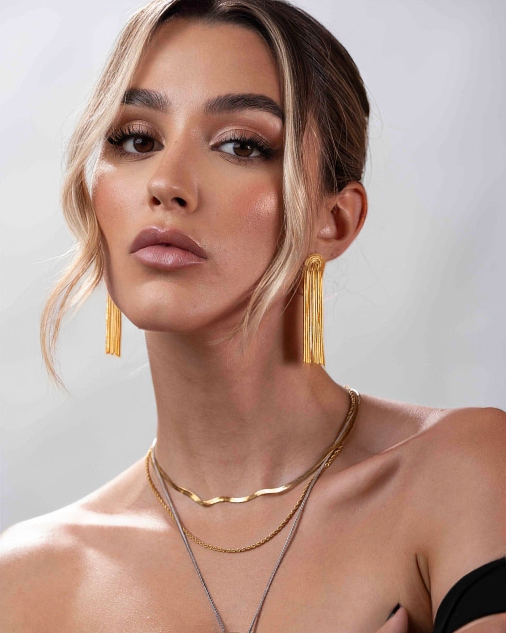 Woman wearing gold earrings and necklaces against a neutral background