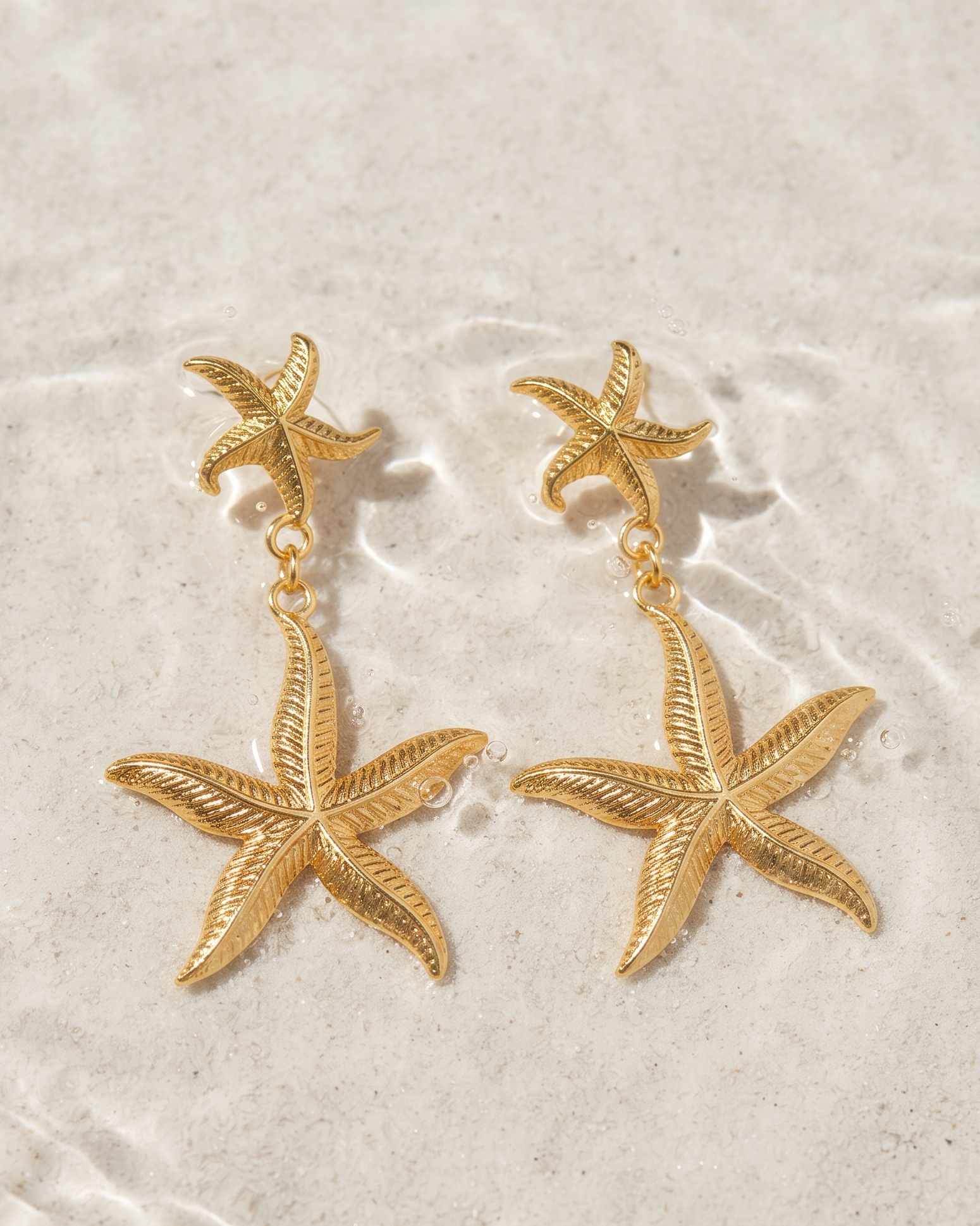 A pair of gold starfish drop earrings displayed on a textured, light beige sandy surface.