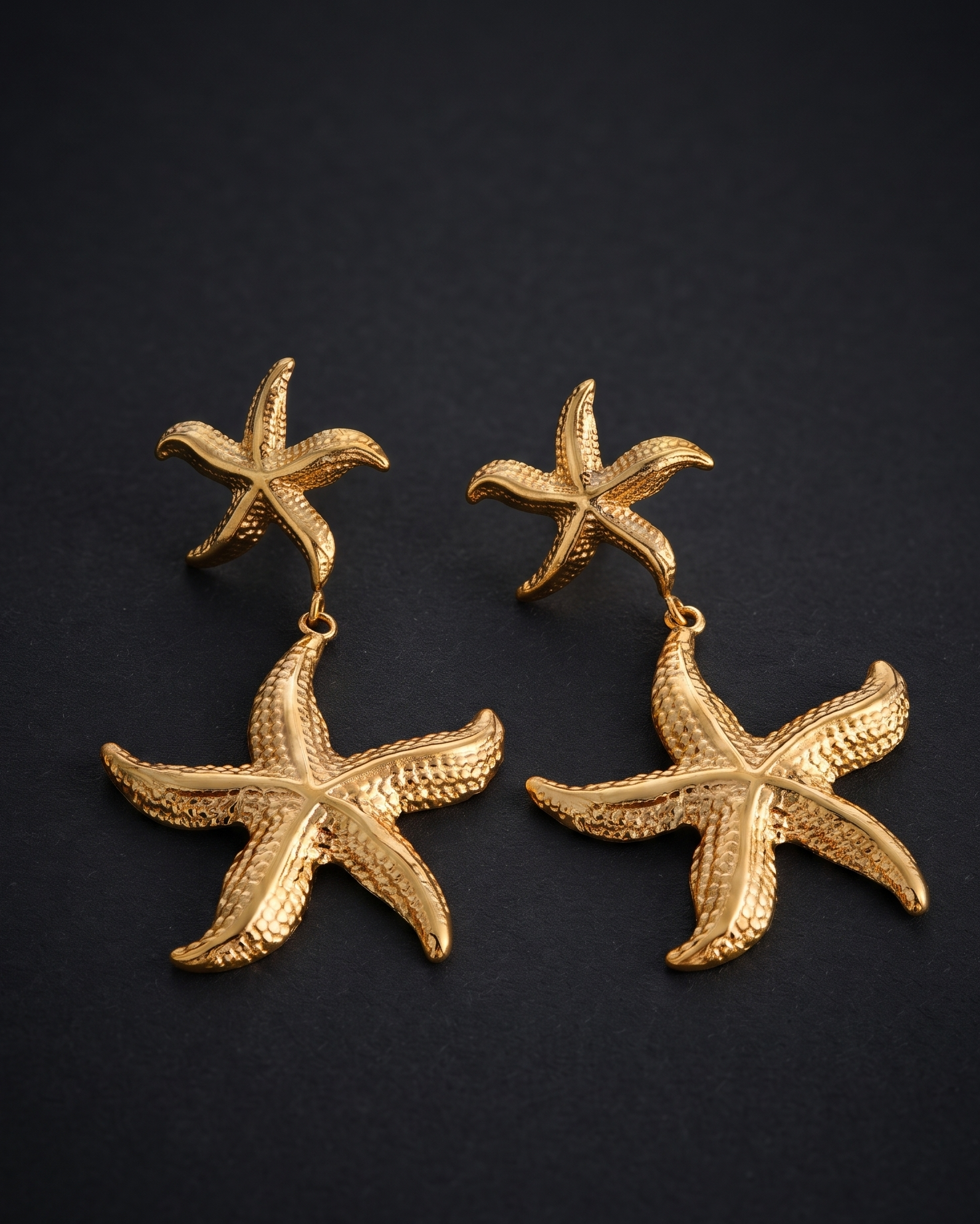 A pair of gold-toned starfish drop earrings on a dark, textured background.