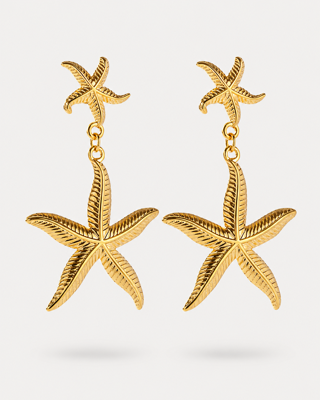 A pair of gold starfish drop earrings with a smaller starfish dangling above a larger textured starfish on each earring, shown against a light gray background.