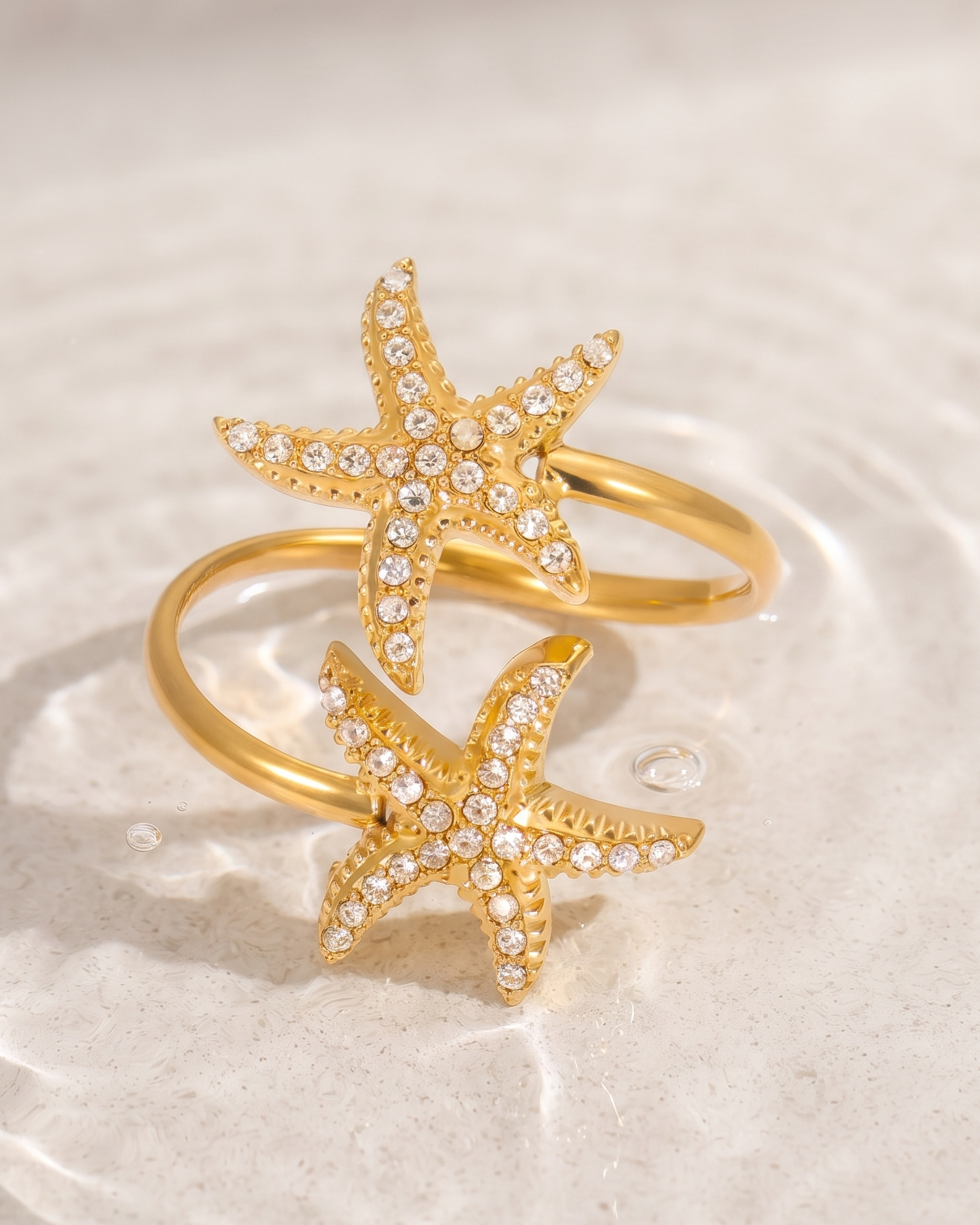 A pair of gold starfish rings adorned with small clear crystals, resting on a light beige textured surface with soft water ripples in the background.