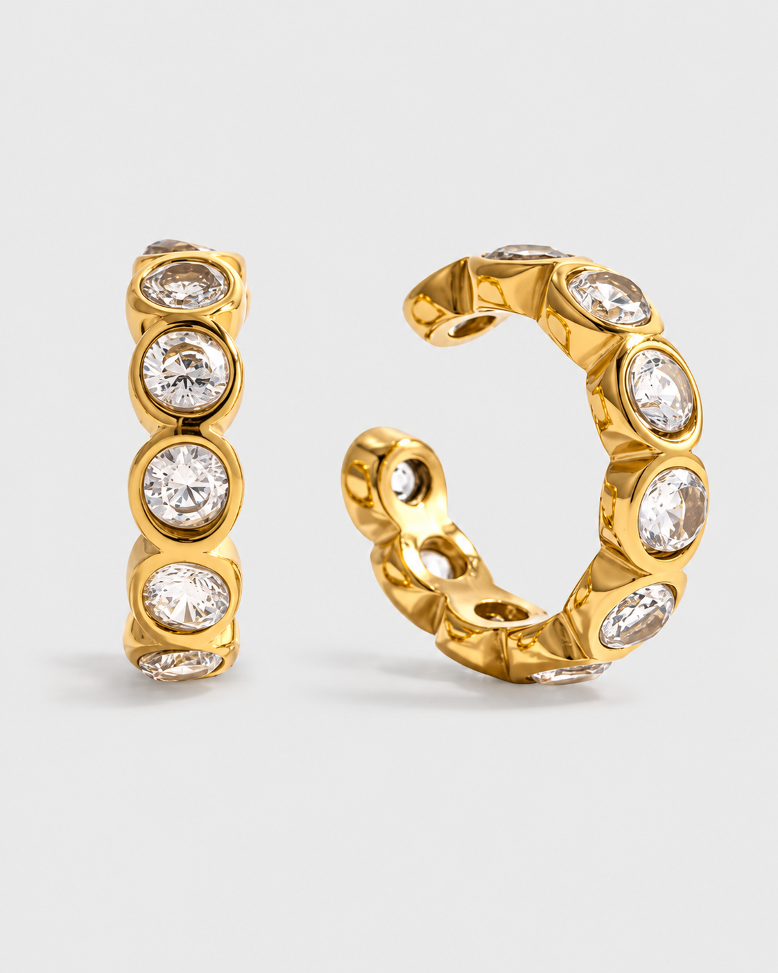 A pair of gold huggie hoop earrings with round clear gemstones, shown on a light gray background.