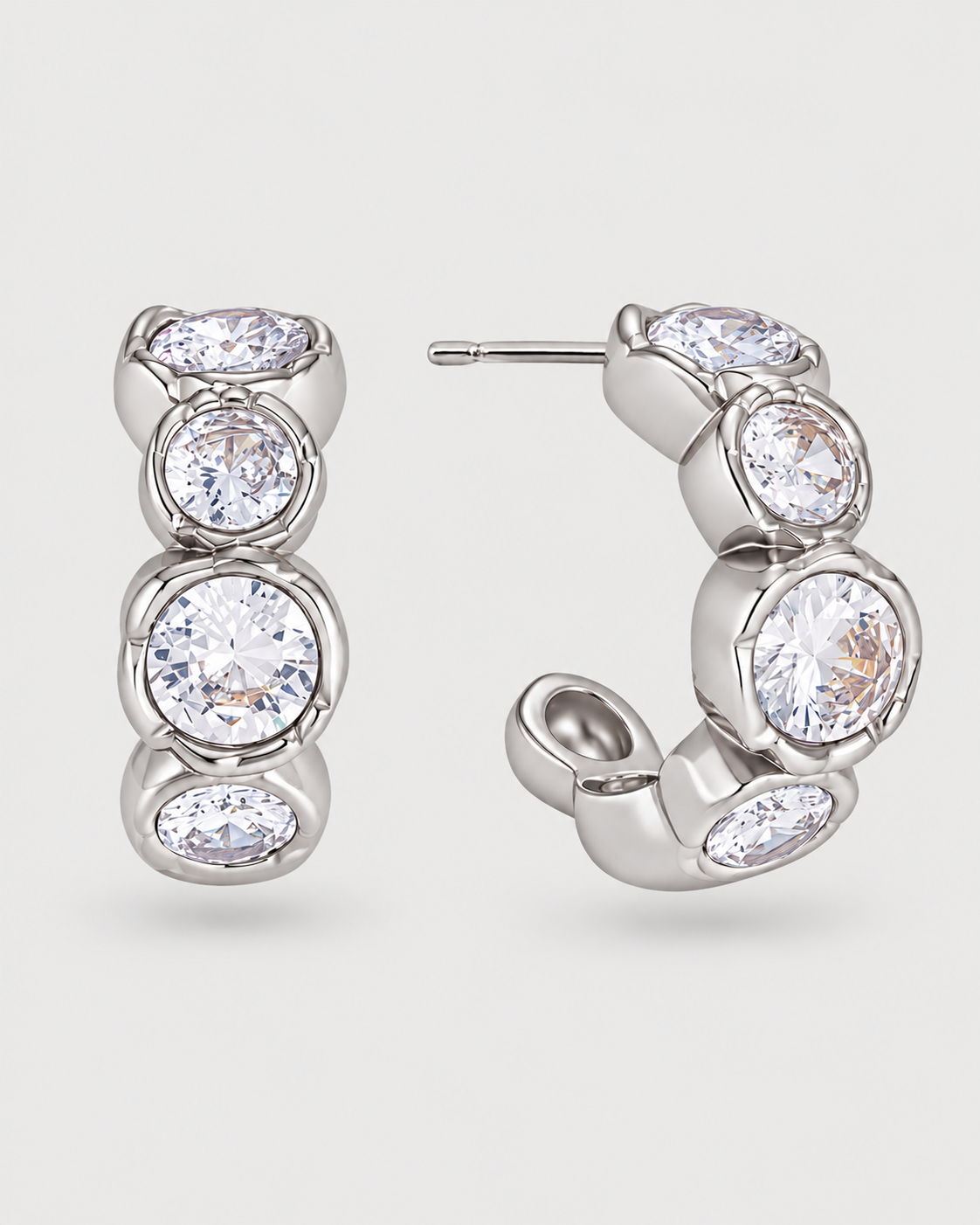 A pair of silver hoop earrings with bezel-set clear oval and round gemstones, displayed on a light gray background.