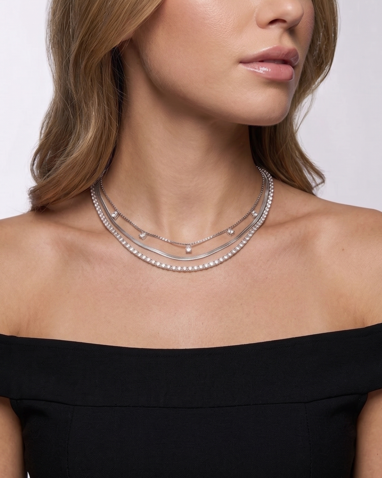 Woman wearing a black off-shoulder top and multiple necklaces on a plain background