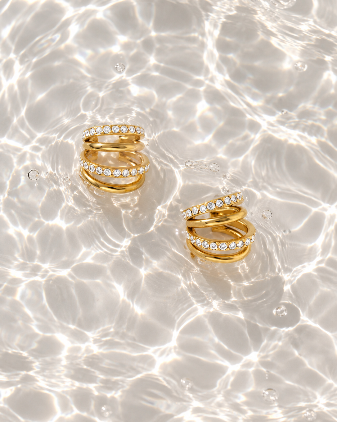 Two gold spiral rings with rows of clear pavé stones, floating on rippling water with light reflections.