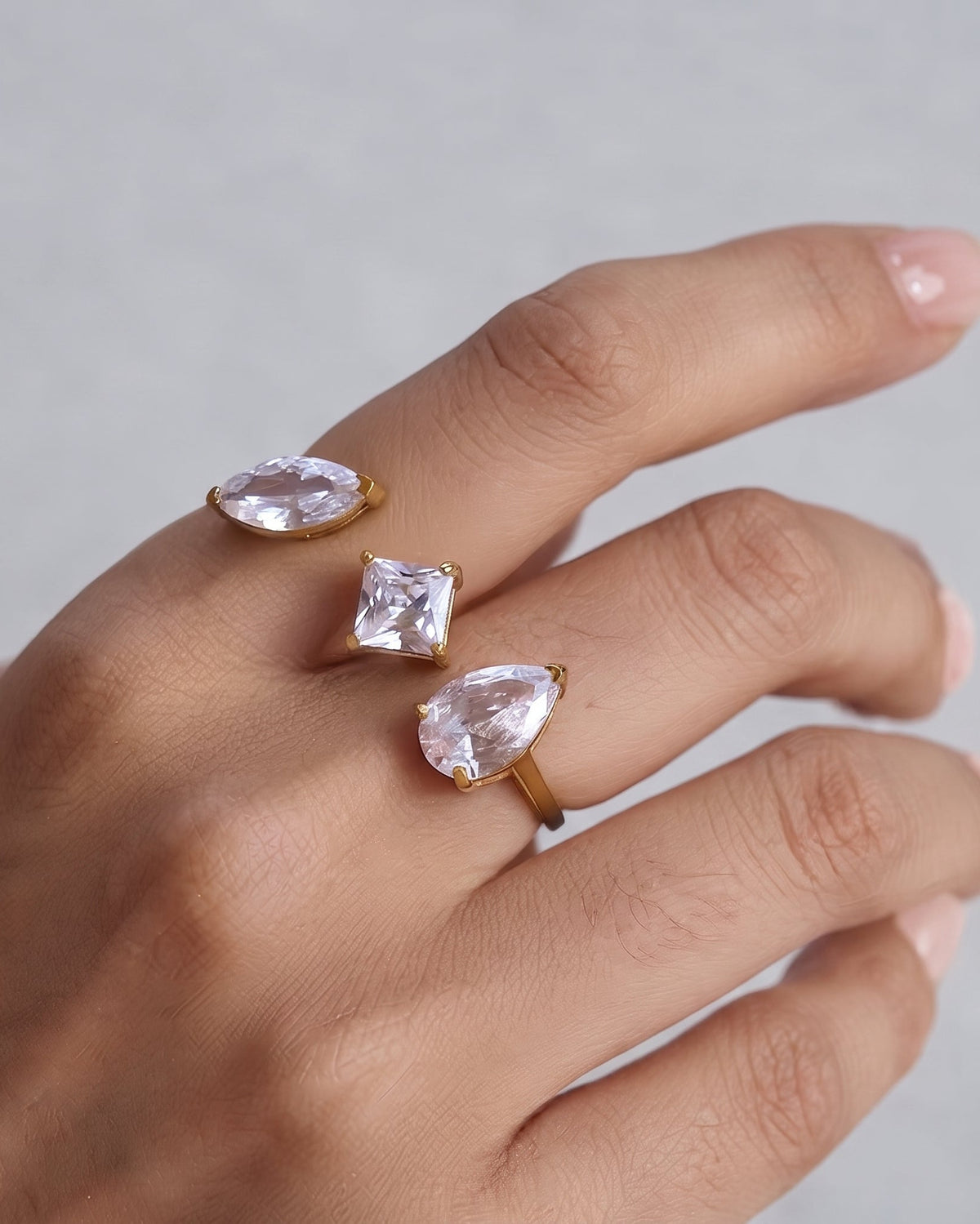 A close-up of a hand wearing three gold rings with clear teardrop and square gemstones against a soft gray background.