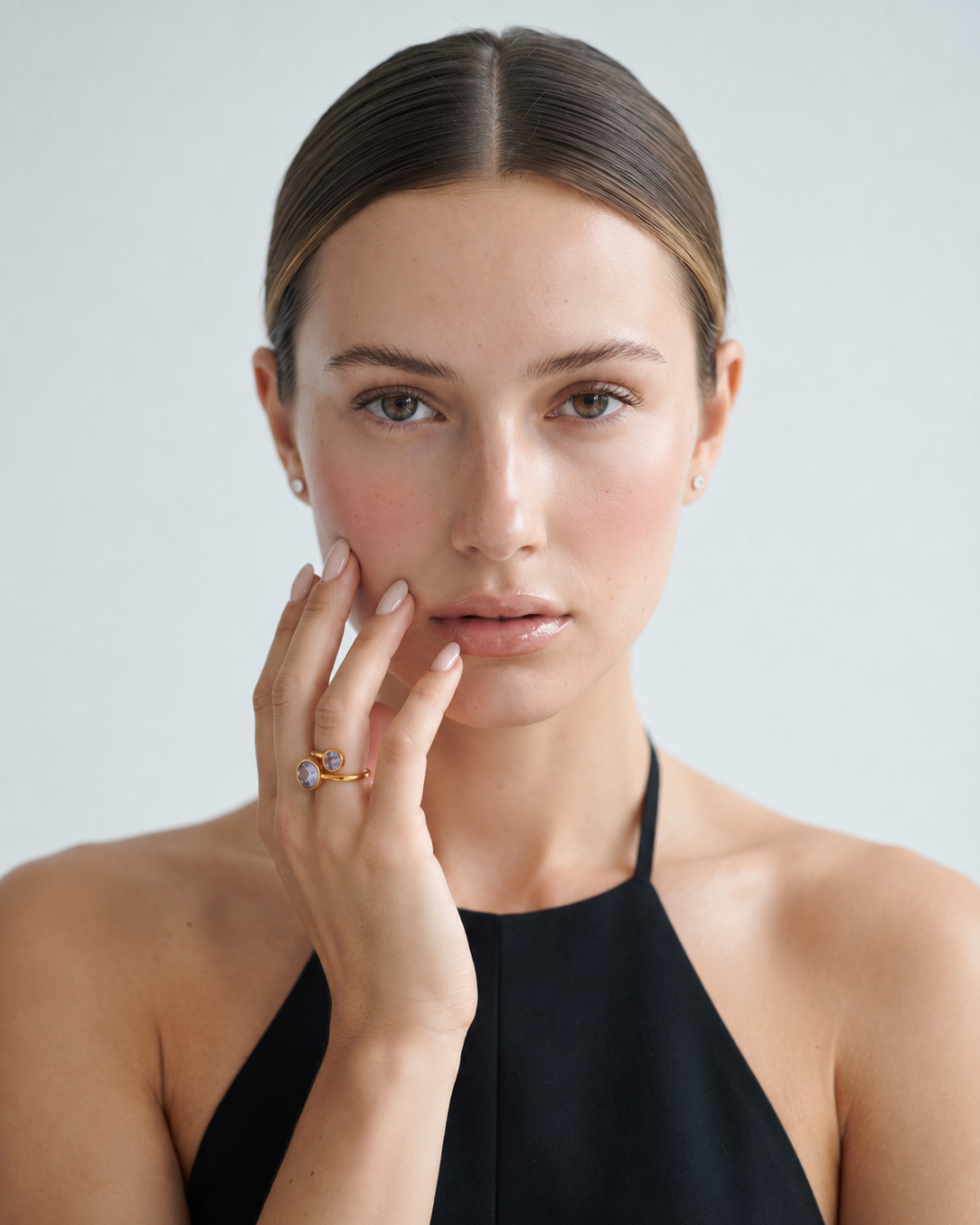 A close-up portrait of a woman wearing a black halter top and a delicate gold ring with a small blue gemstone on her finger, set against a light gray background.