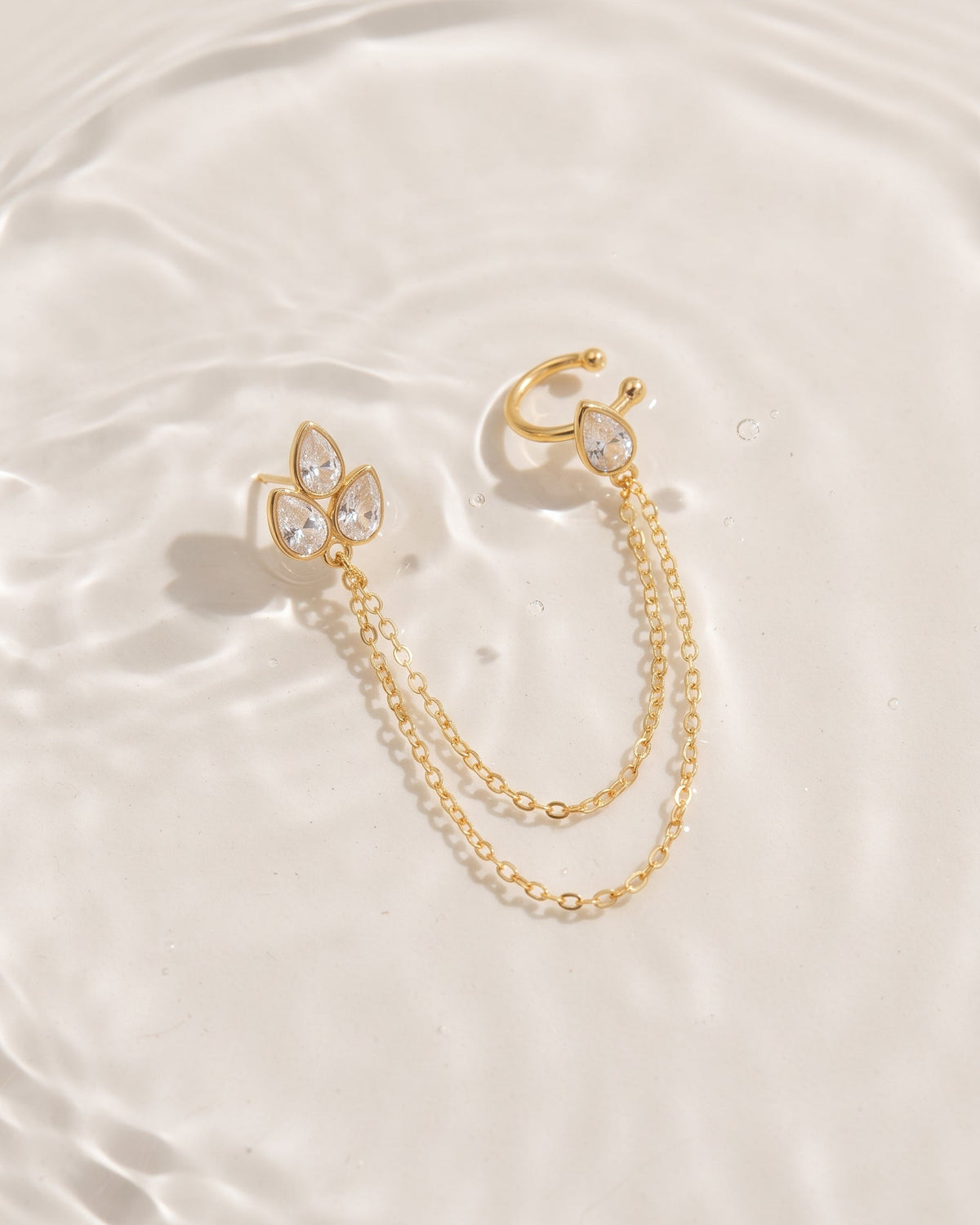 A pair of gold ear cuffs with teardrop crystal details and double chain drops, displayed on a clean, textured white surface.