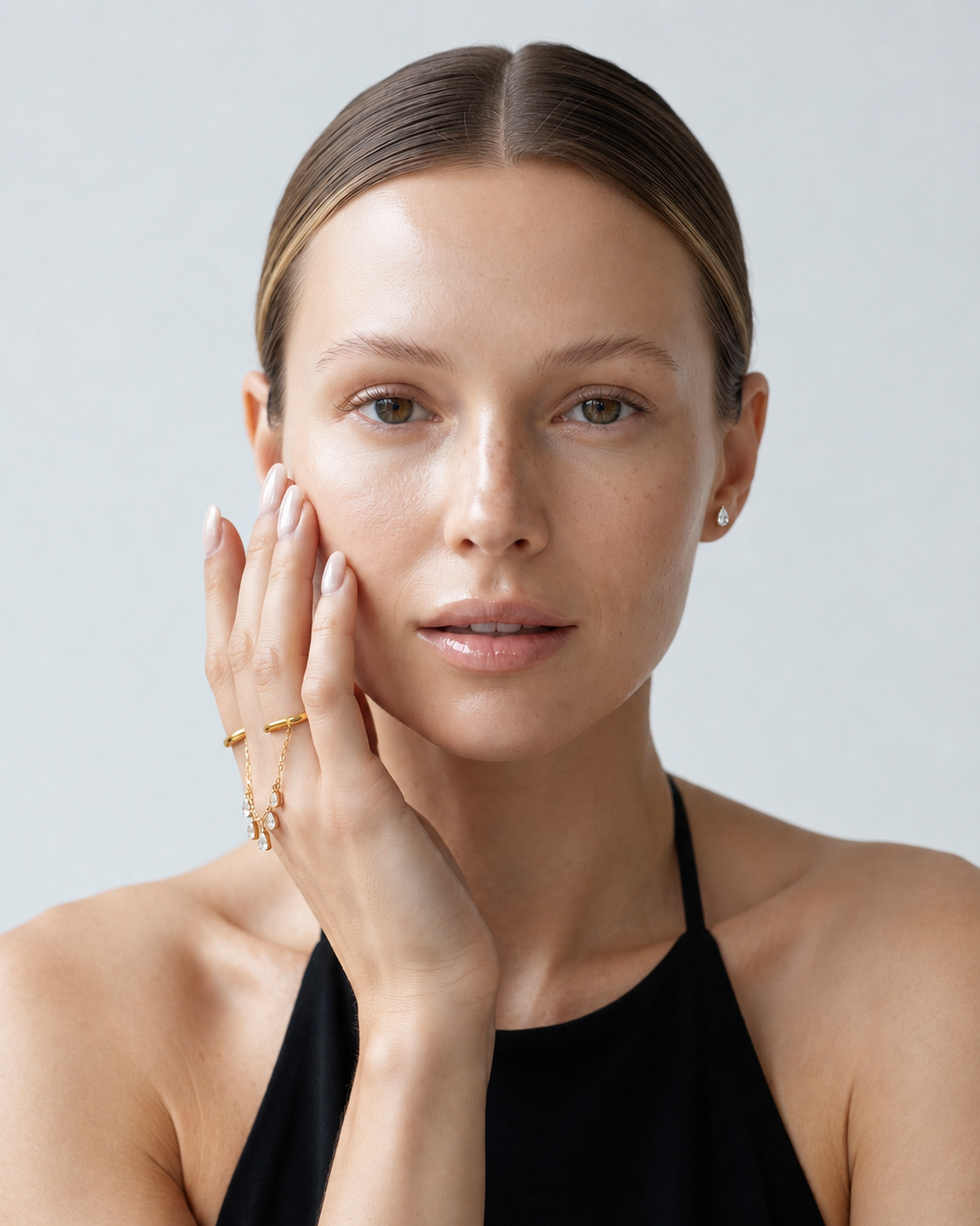 A close-up portrait of a woman with her hand resting on her cheek, wearing delicate gold rings and a small stud earring against a light gray background.