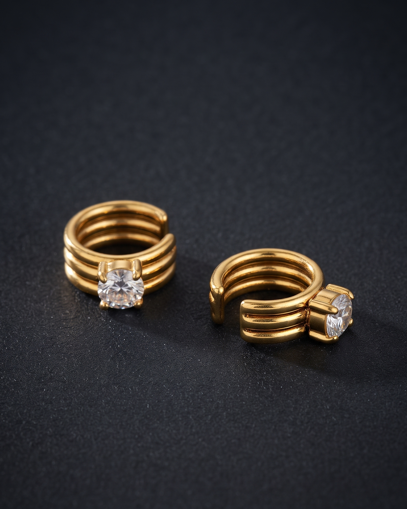A pair of gold-toned open hoop rings with a single clear gemstone on each ring, displayed on a dark textured background.