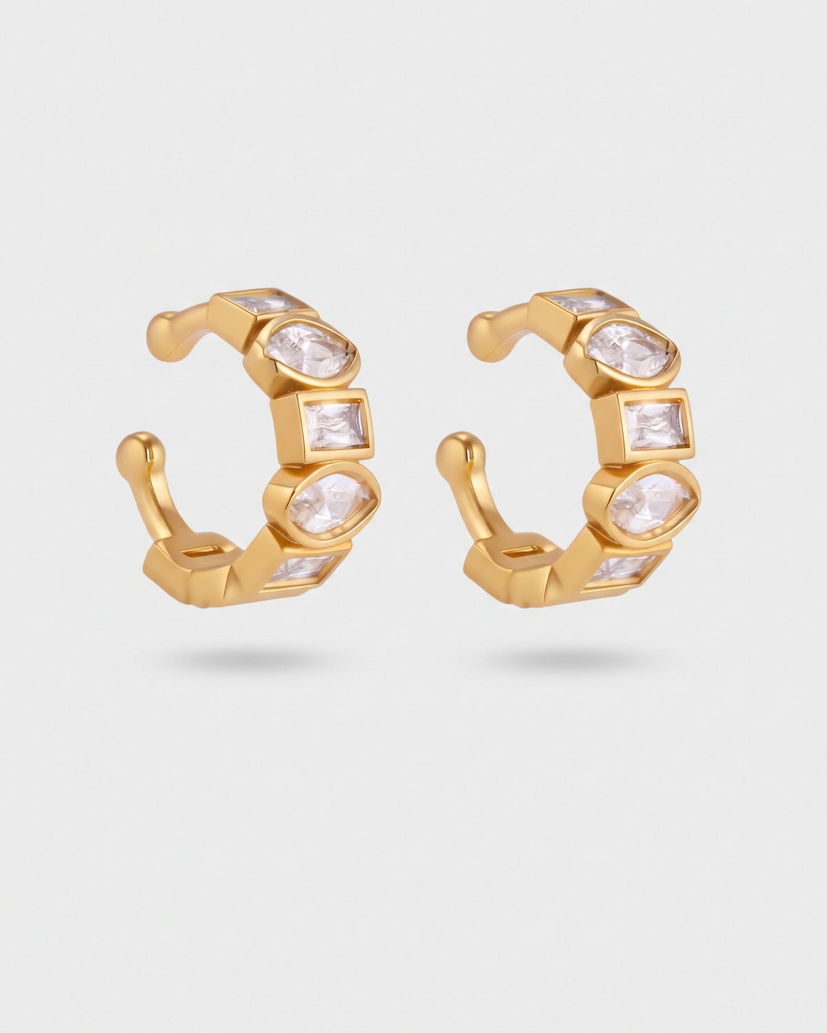 A pair of gold huggie hoop earrings with clear faceted gemstones, displayed on a light gray background.