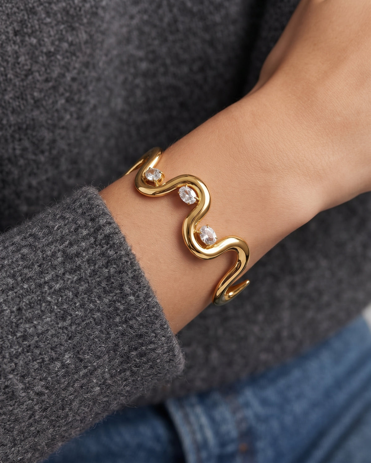 A gold wavy cuff bracelet with three clear gemstones, worn on a person's wrist over a gray knit sweater and blue jeans.