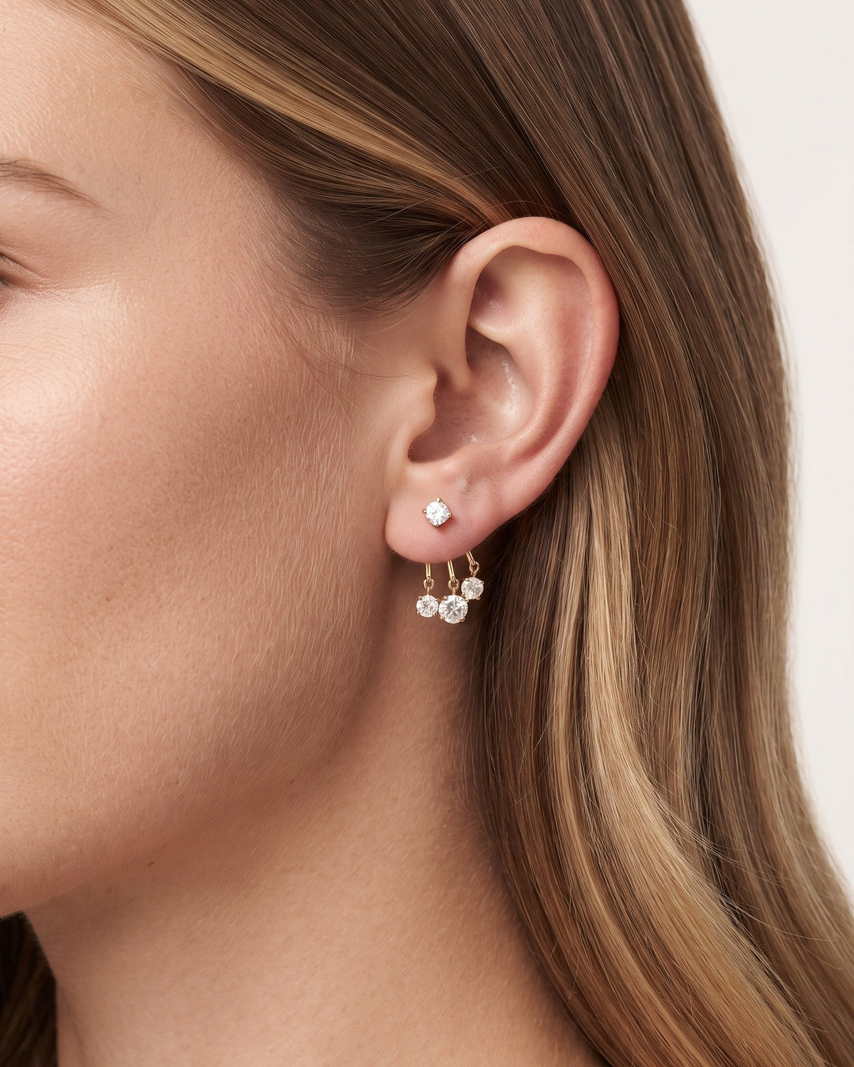 A close-up side profile of a woman's ear wearing a delicate gold dangle earring with small clear gemstones, against a soft light beige background.