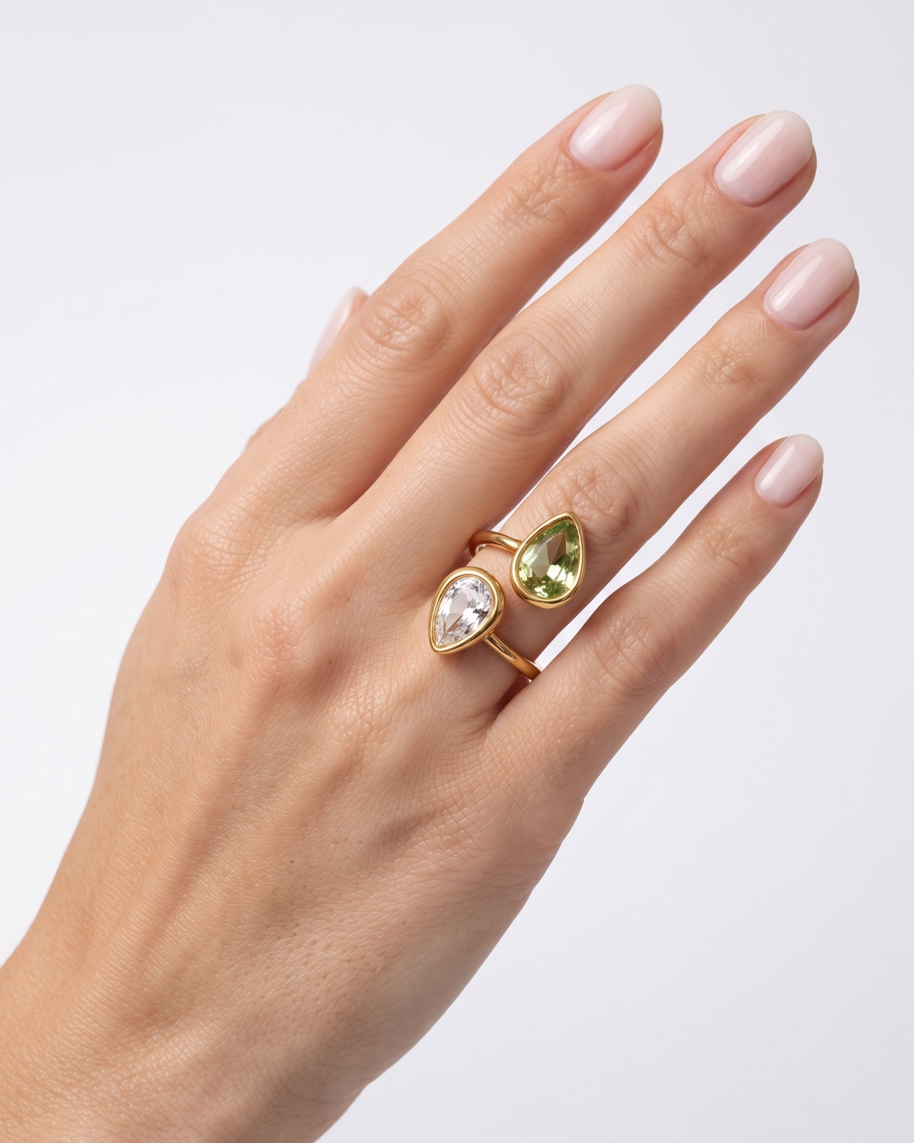 A close-up of a hand wearing two gold rings with teardrop-shaped gemstones, one clear and one light green, against a light gray background.