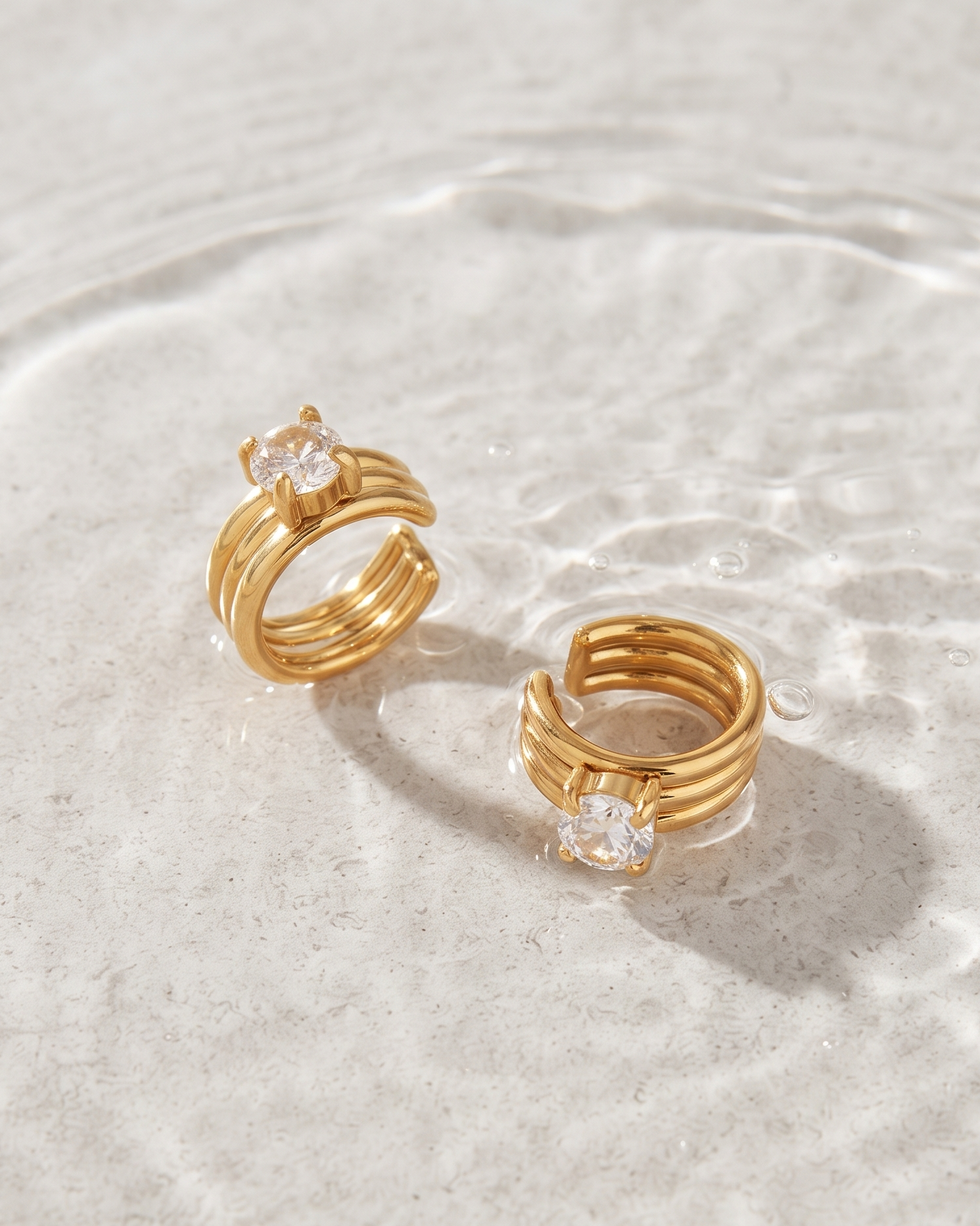 A pair of gold-plated multi-band rings with clear round-cut gemstones, displayed on a textured light gray surface with water ripples.