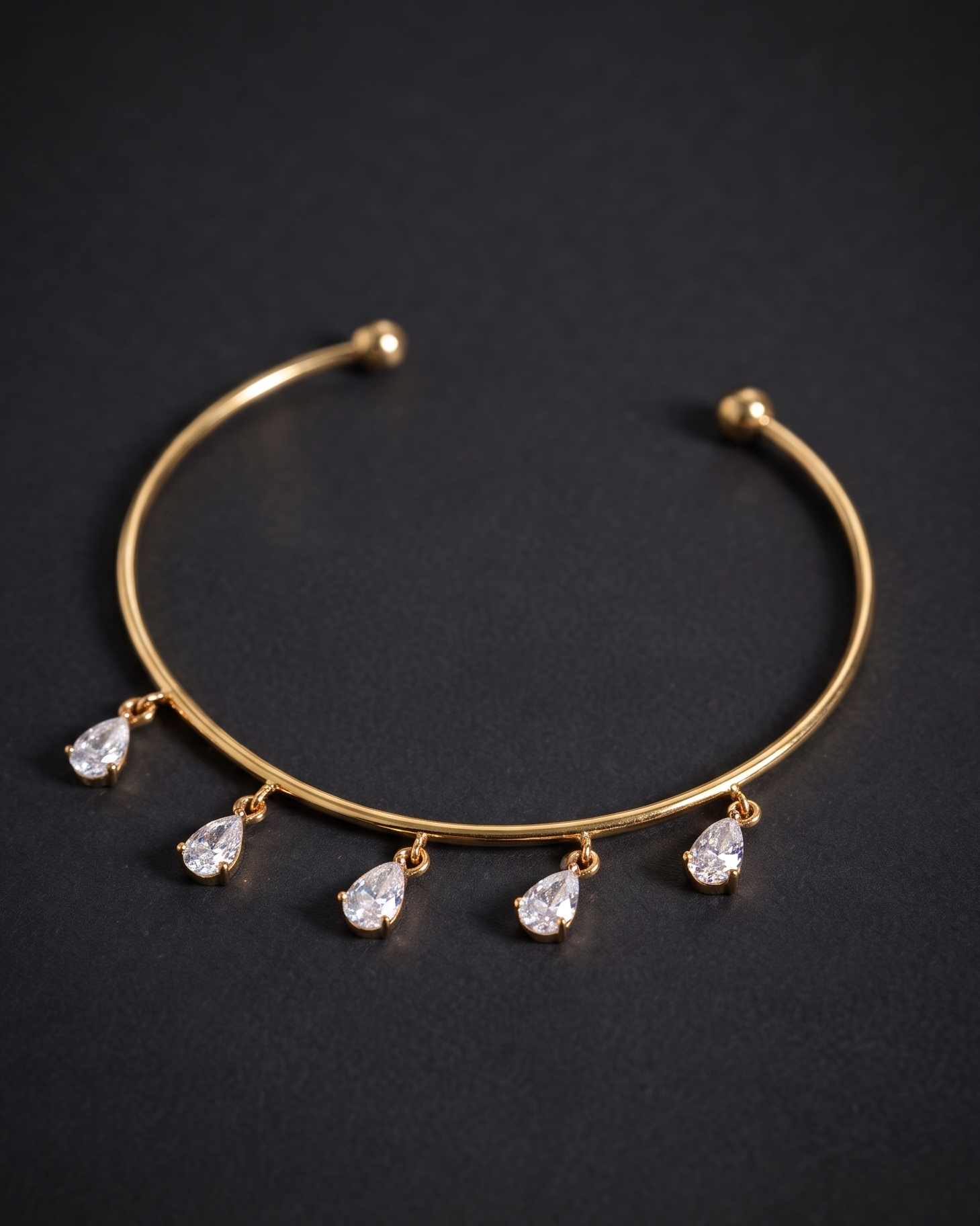 A gold-toned open cuff bracelet with five dangling teardrop-shaped clear gemstones, photographed on a dark textured background.