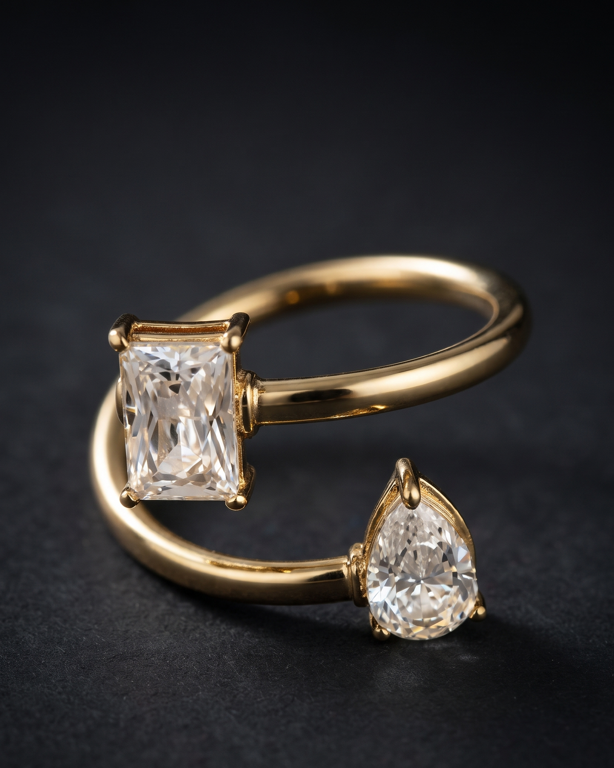 A gold open ring necklace with two clear gemstones, one rectangular and one pear-shaped, set against a dark textured background.
