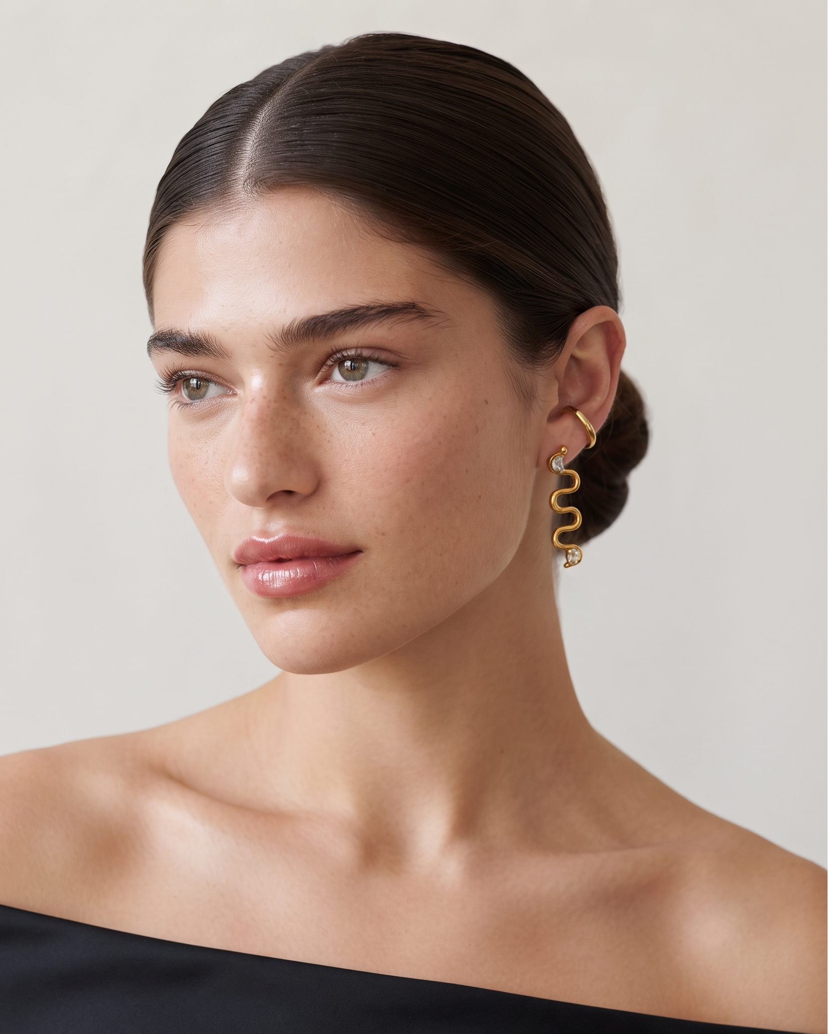 A close-up portrait of a woman with dark hair pulled back, wearing a gold spiral drop earring. She is wearing a black off-the-shoulder top against a plain, light beige background.