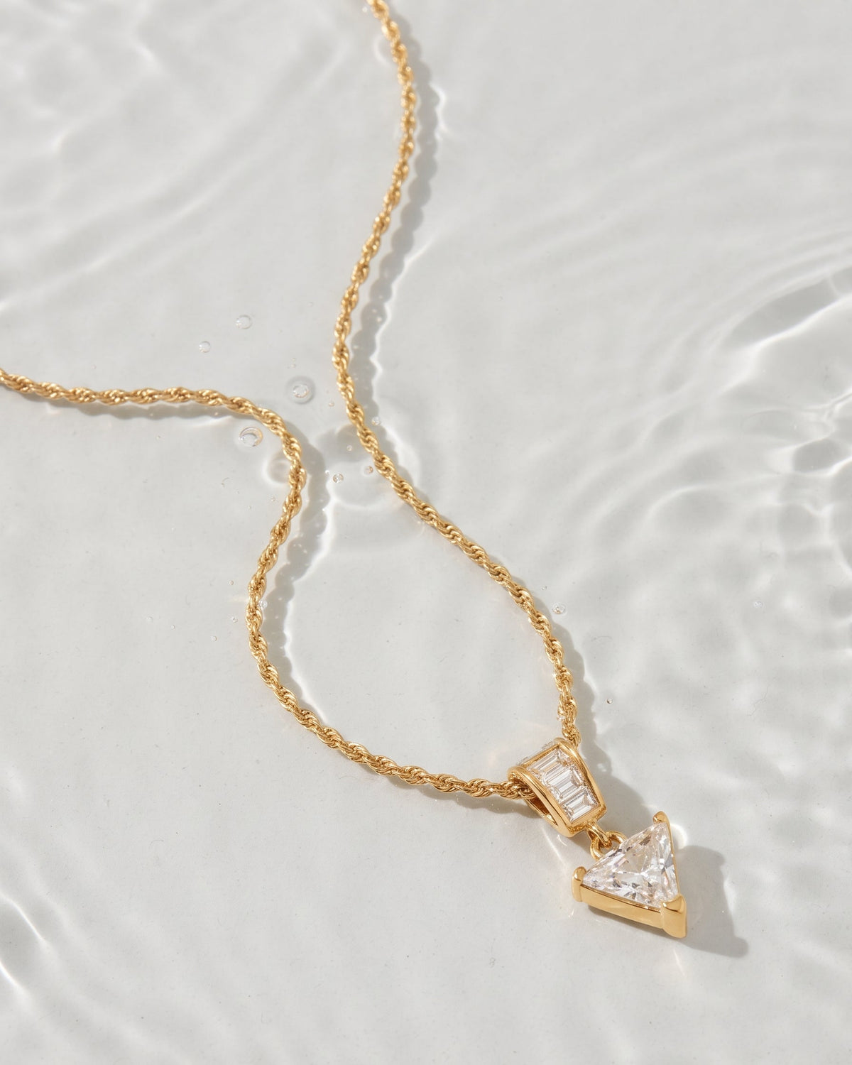 A gold chain necklace with a triangular pendant featuring clear stones, photographed on a glossy white surface with soft water ripples.