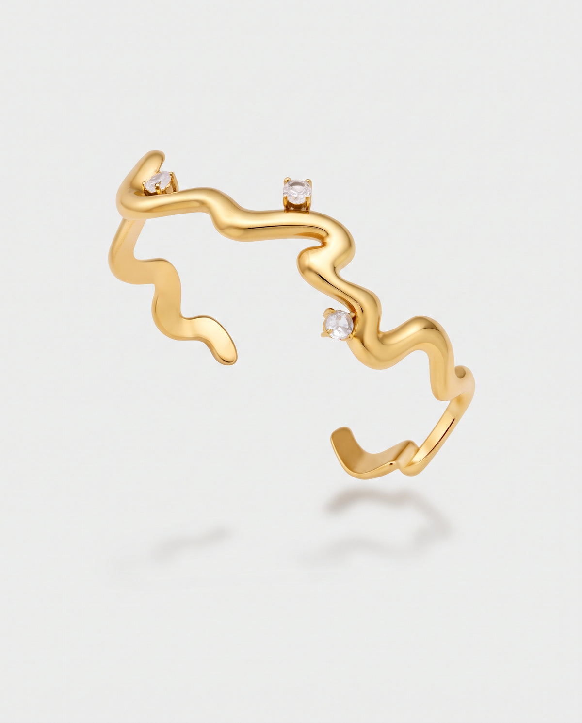 A gold open cuff ring with a wavy, fluid organic line design and three small clear gemstone accents, shown on a light gray background.