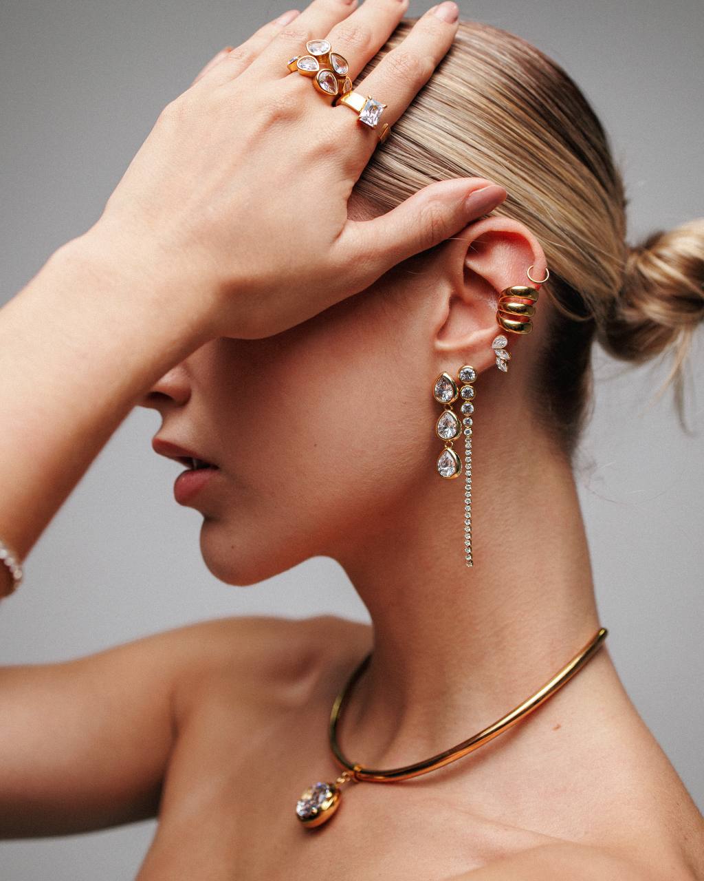 A close-up lifestyle shot of a model wearing elegant gold jewelry, including a twisted gold choker necklace with a teardrop pendant, a matching gold hoop earring, and gold rings on her fingers.