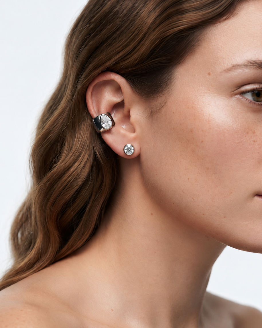 A close-up of a woman's ear wearing a silver-toned ear cuff with a clear gemstone, alongside a matching small stud earring, against a light gray background.