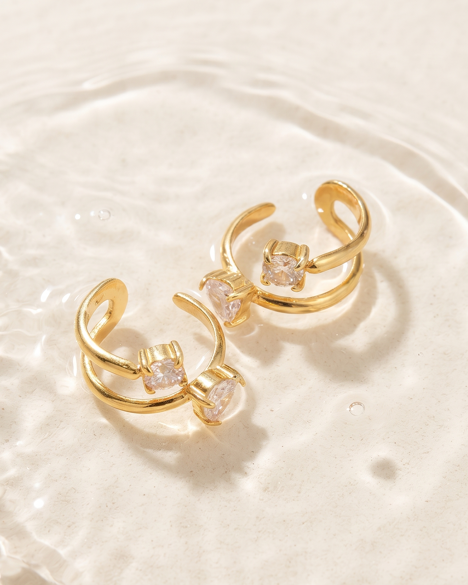 A pair of gold-toned open hoop earrings with clear gemstones, resting on a textured beige surface with water ripples.