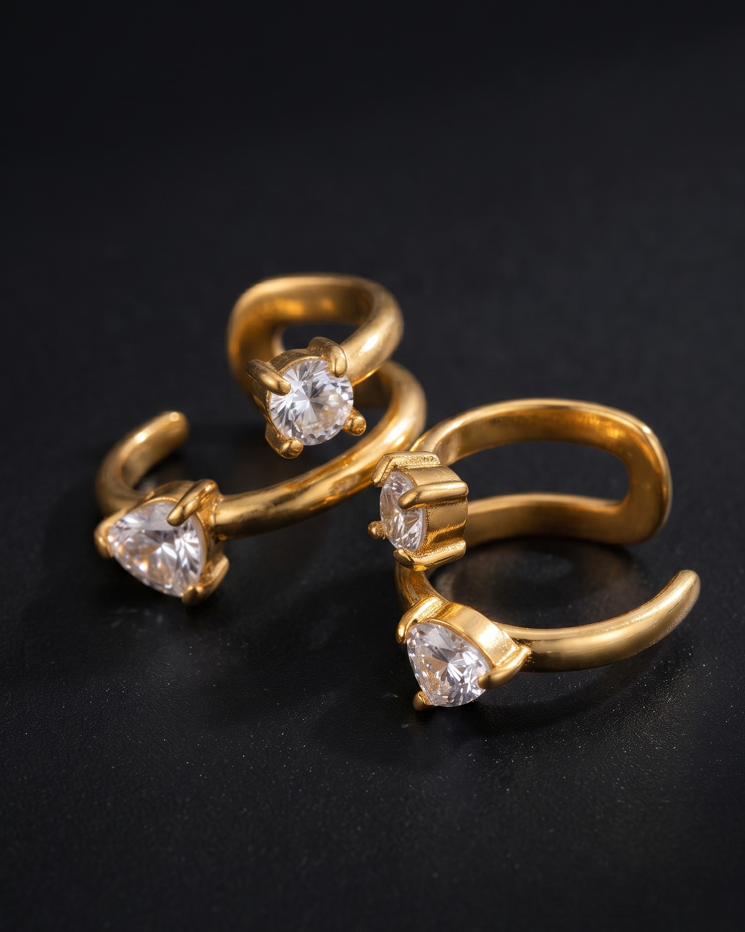 Three gold-toned open ring earrings with heart-shaped clear gemstones, arranged on a dark black background.