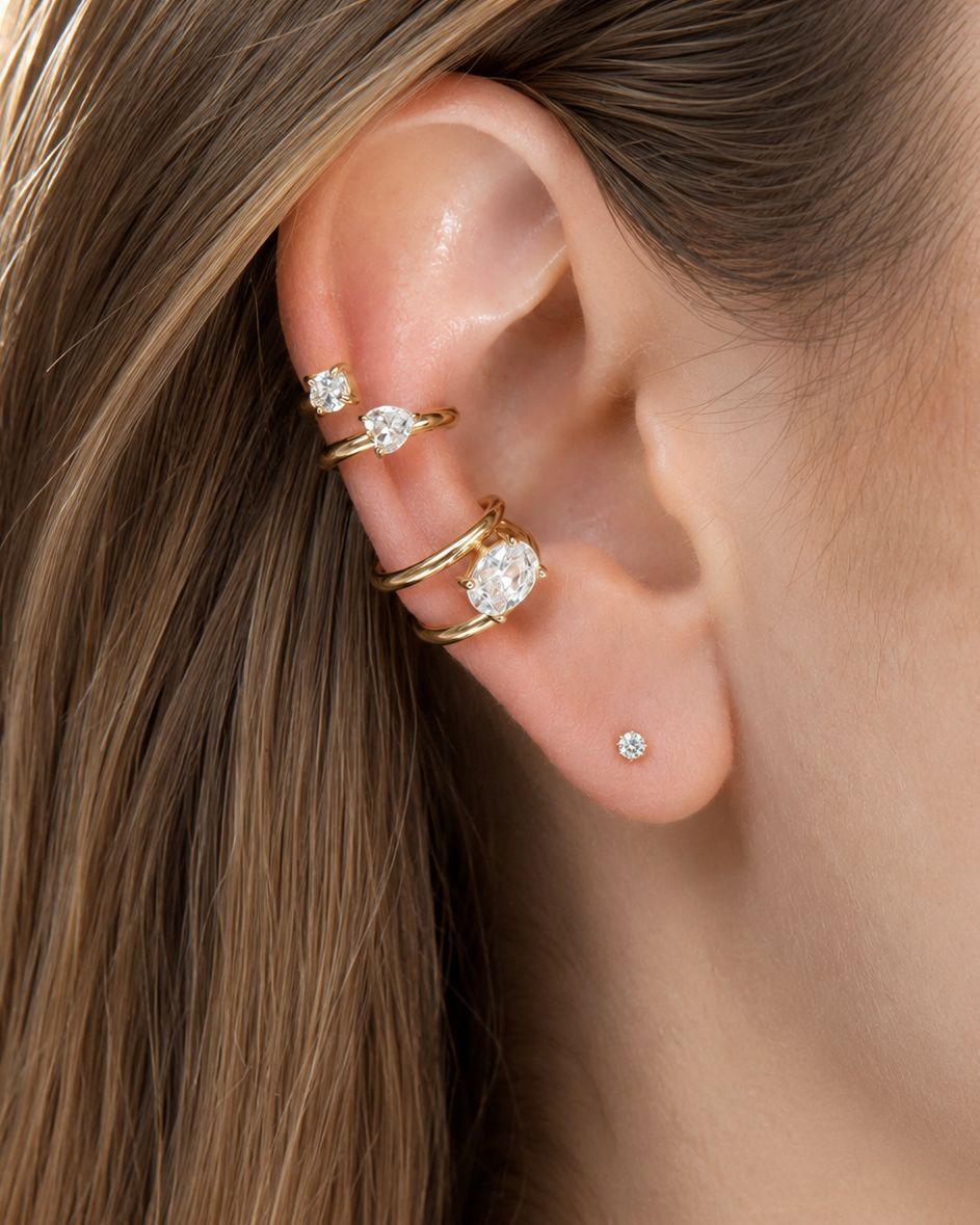 A close-up of a person's ear showcasing a gold ear cuff set with clear gemstones, along with a small stud earring.