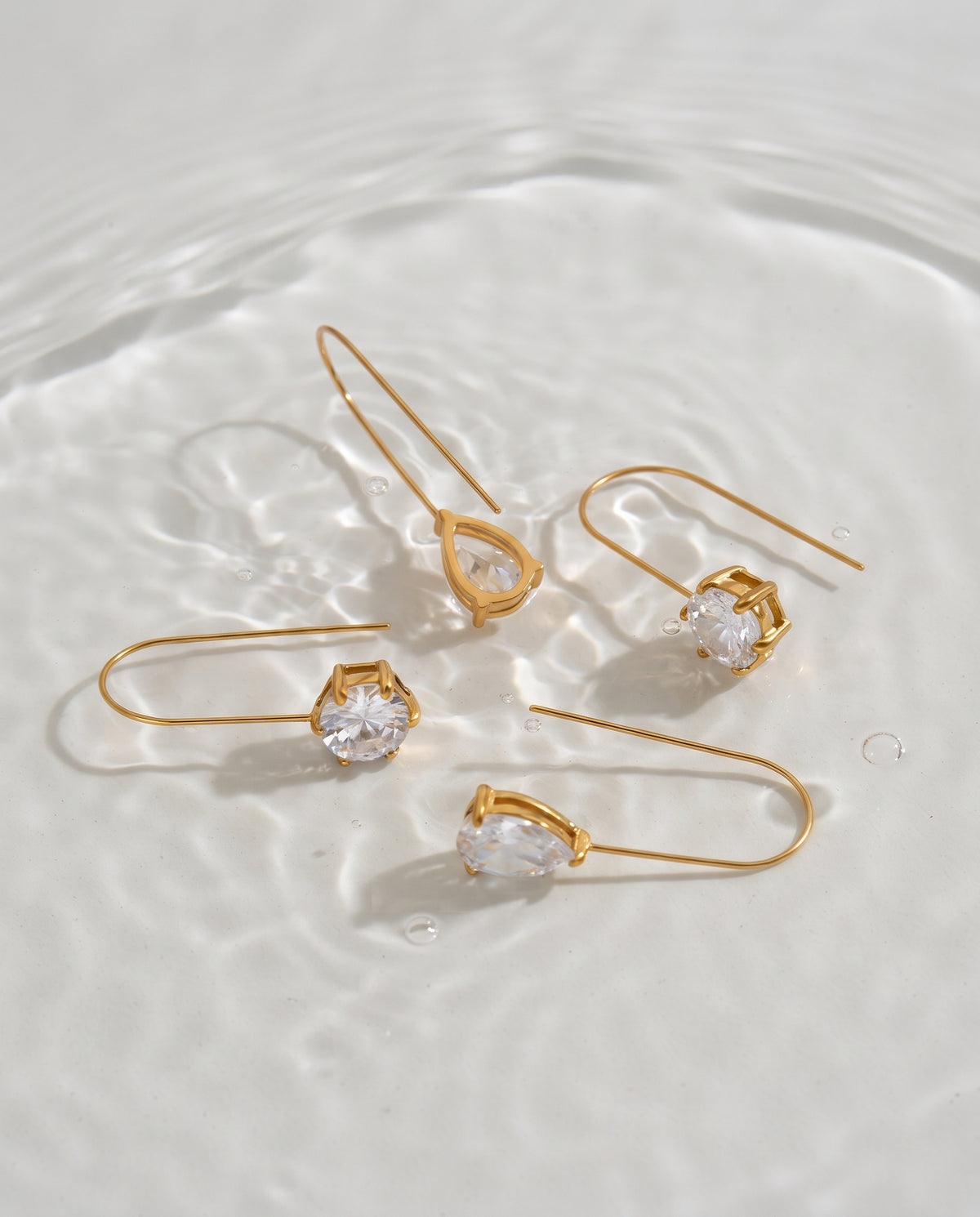 Four gold-tone drop earrings with clear teardrop-shaped gemstones, arranged on a glossy white surface with soft water ripples.