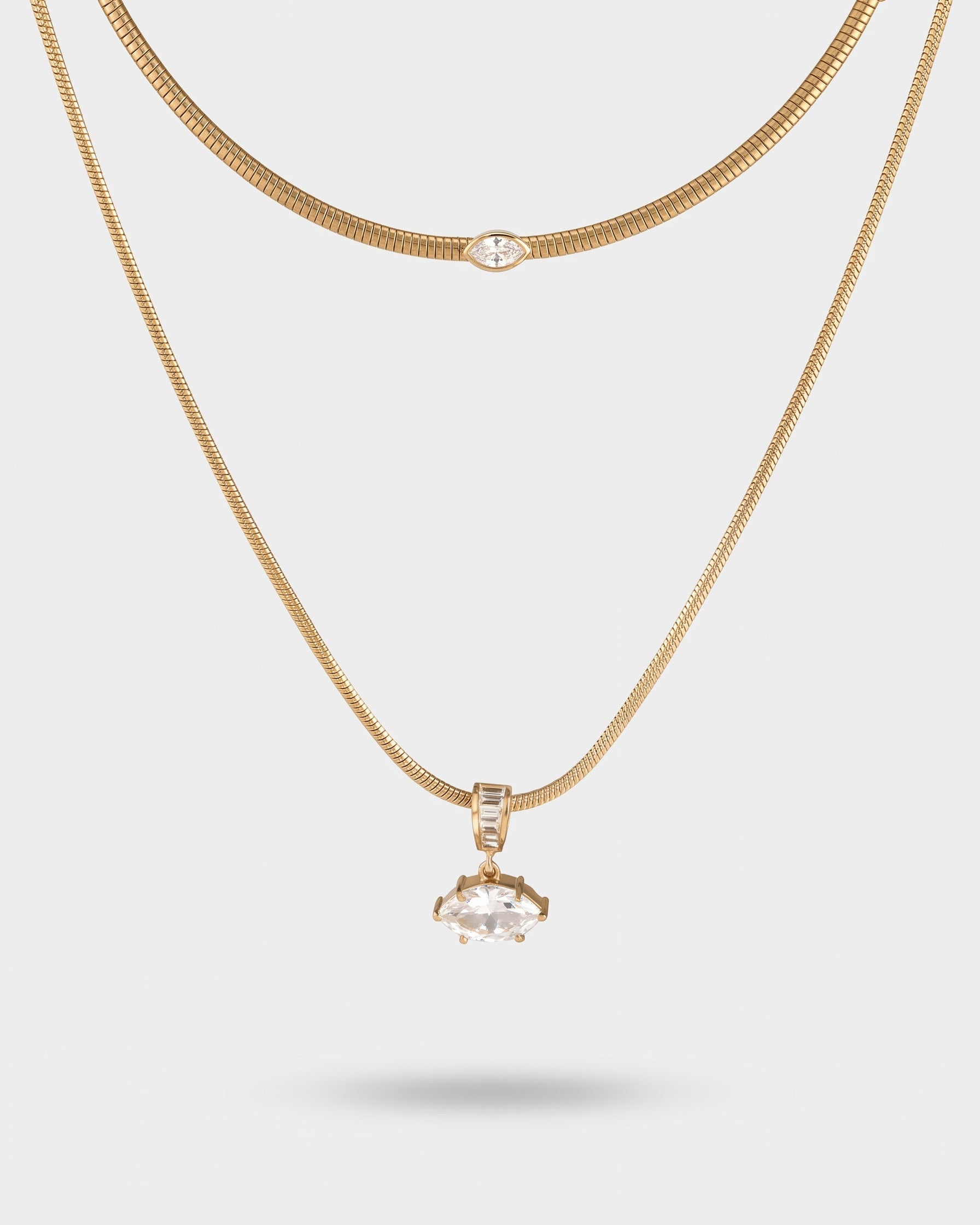 A gold layered necklace set featuring a delicate chain with a small marquise-cut clear stone and a longer chain with a larger marquise-cut stone pendant, displayed on a light gray background.