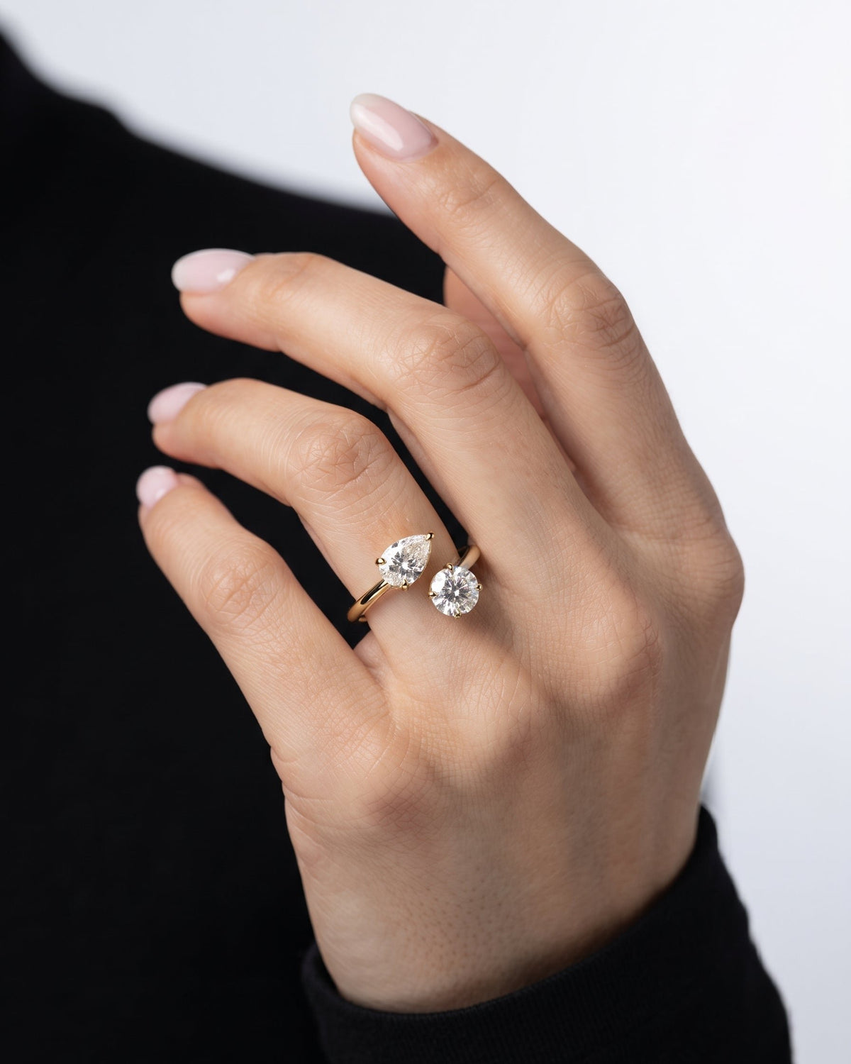 A close-up of a hand wearing two gold rings with clear gemstones, set against a black top and a light gray background.
