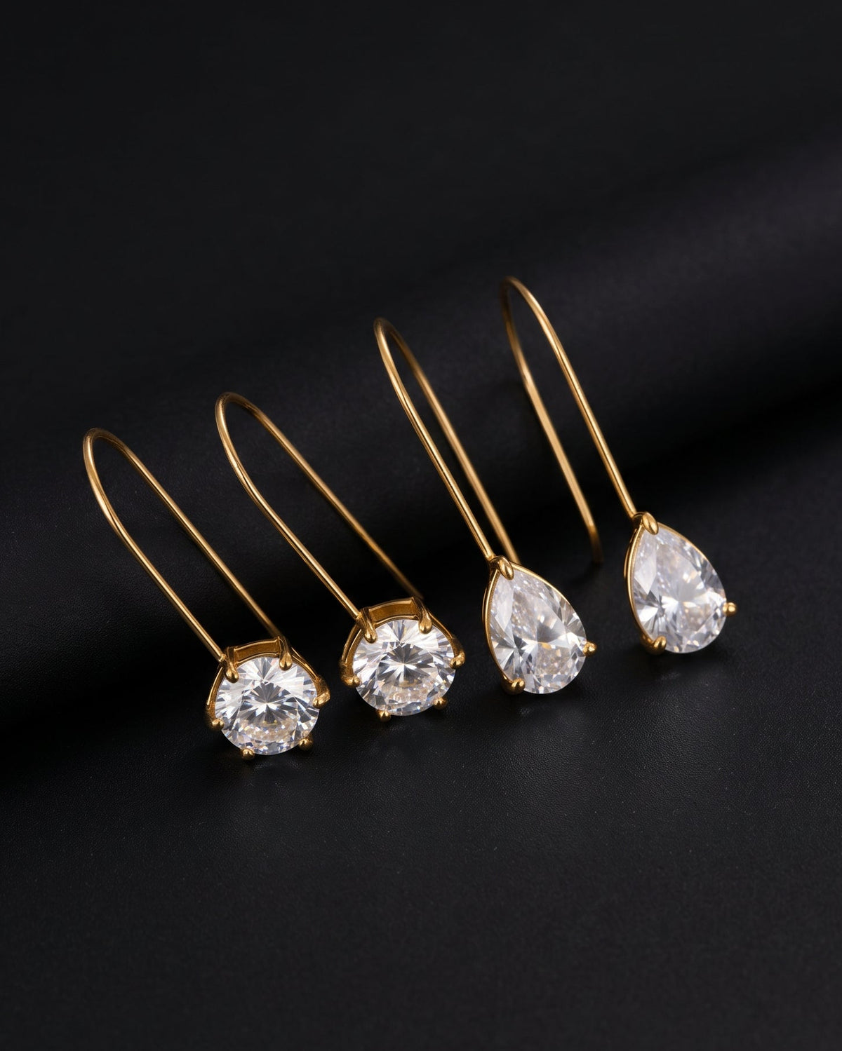 Four gold-tone drop earrings with clear teardrop and round gemstones, arranged diagonally on a dark black background.