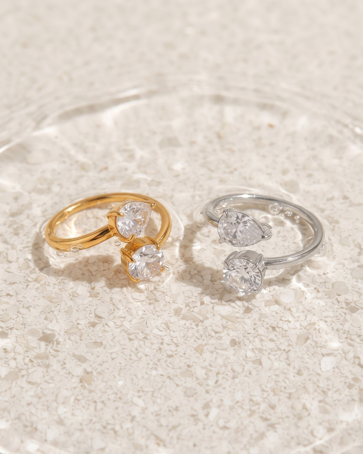 Two rings displayed on a light stone surface: one gold ring with a marquise-cut clear gemstone and one silver ring with a round clear gemstone, both with smaller clear stones along the band.