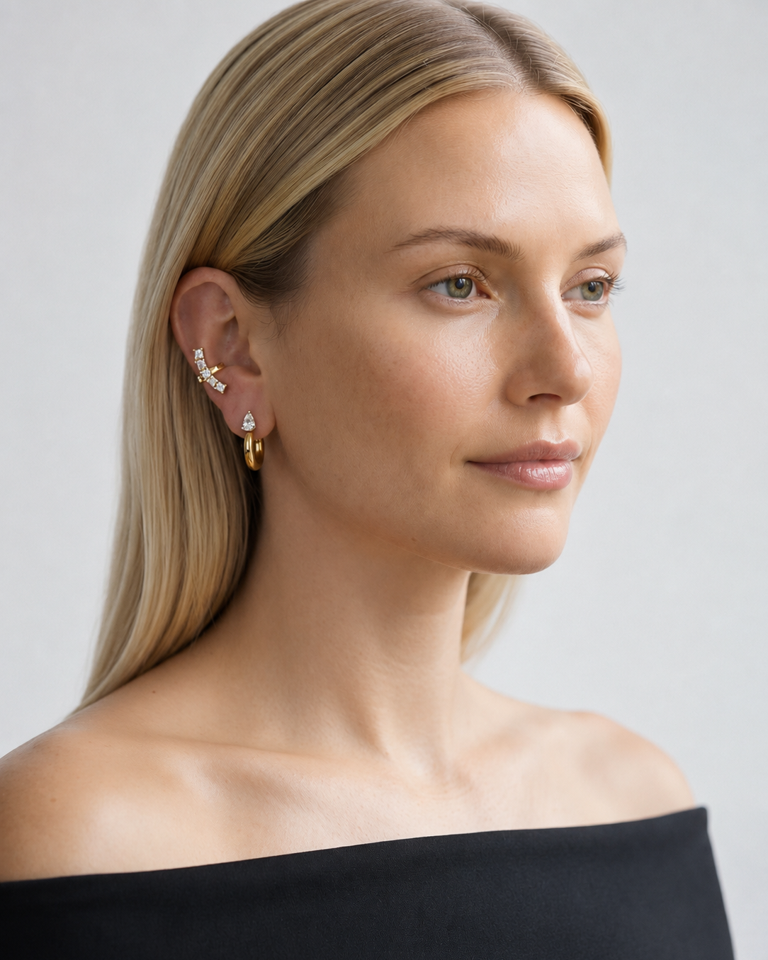 A close-up of a blonde woman wearing a gold ear cuff with small clear stones and a gold hoop earring, shown against a light gray background.