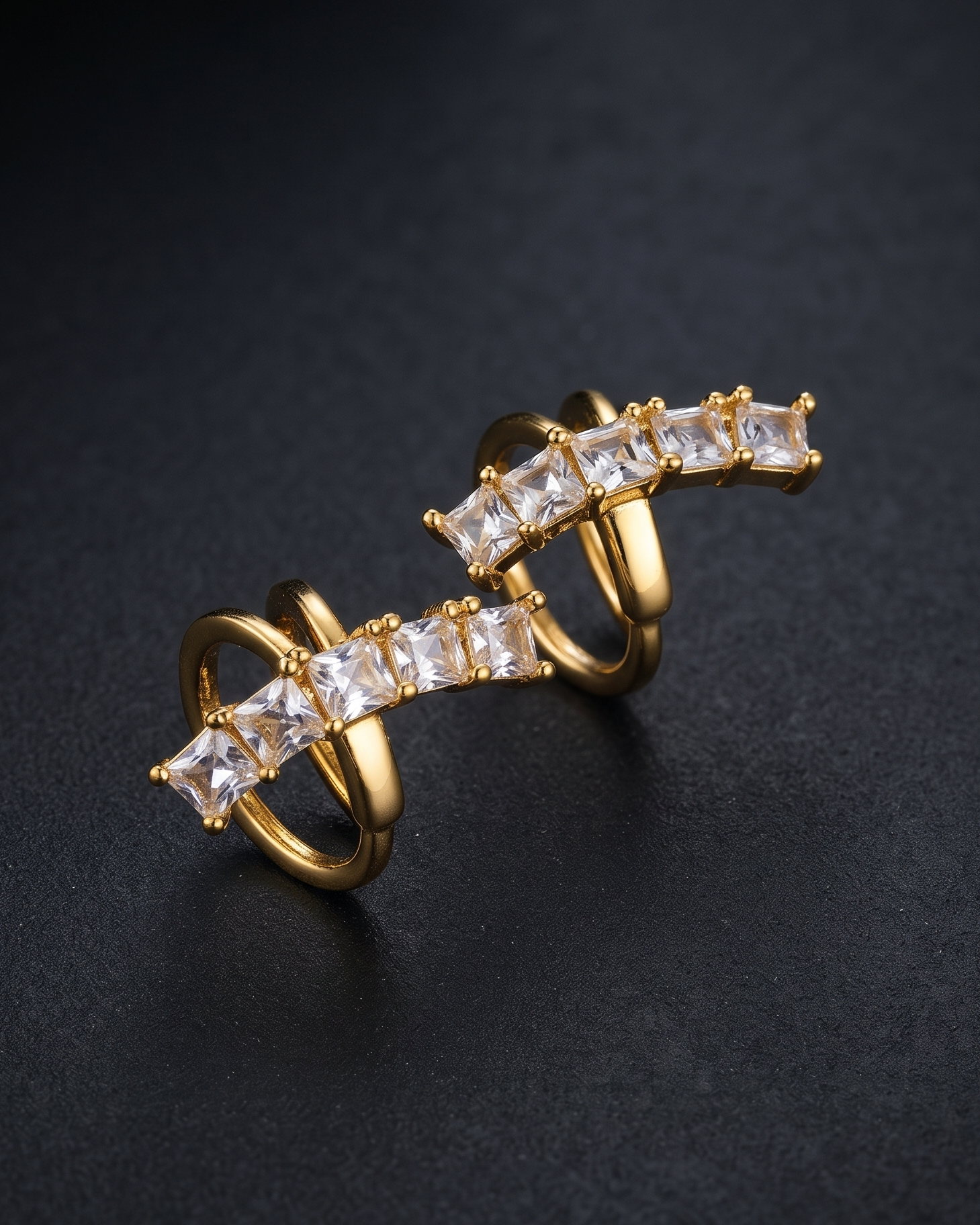 A pair of gold-plated ear cuffs decorated with clear rectangular baguette crystals, displayed on a dark textured background.